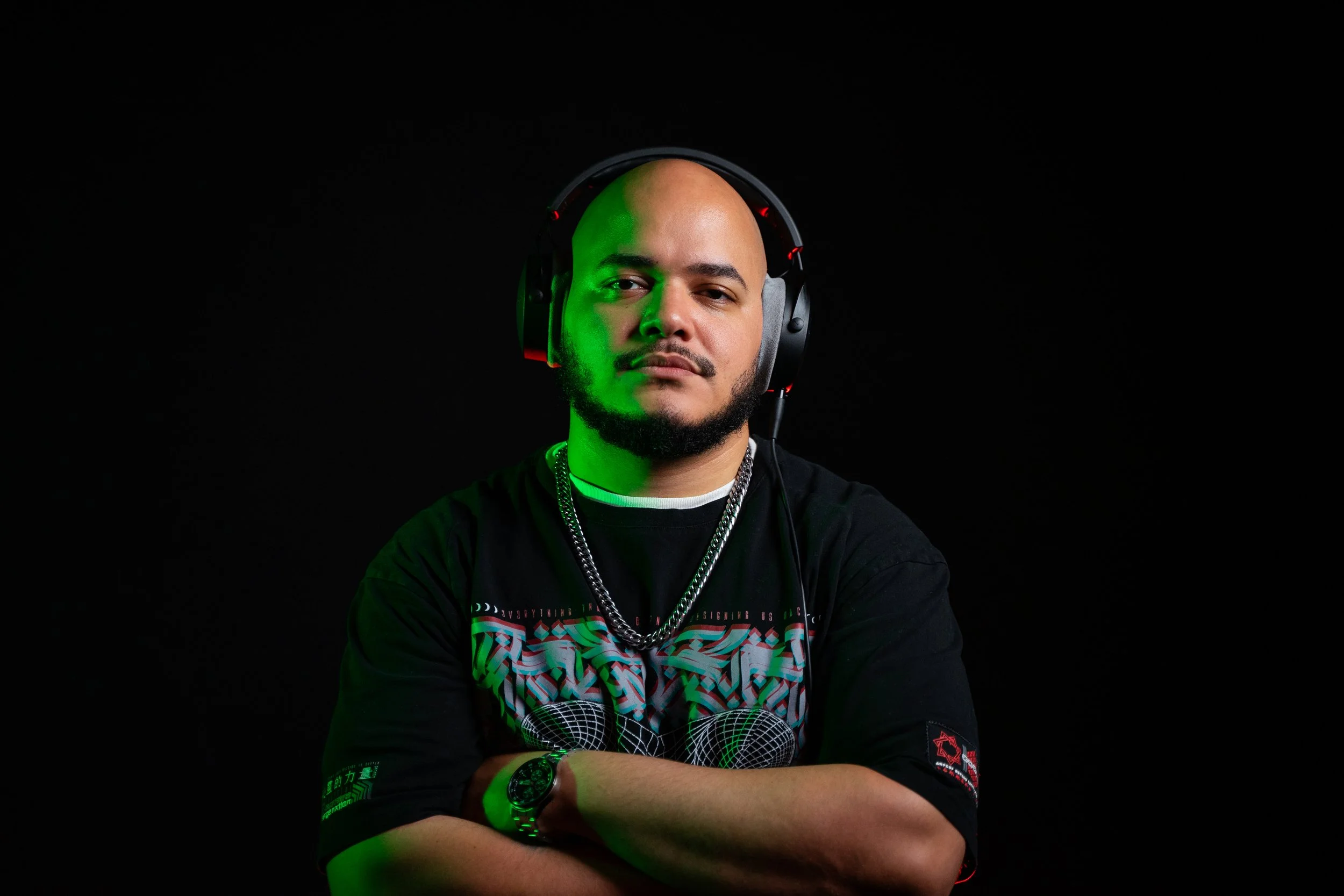 A man with a bald head and beard wearing headphones, a black graphic T-shirt, a watch, and a chain necklace, standing against a black background.