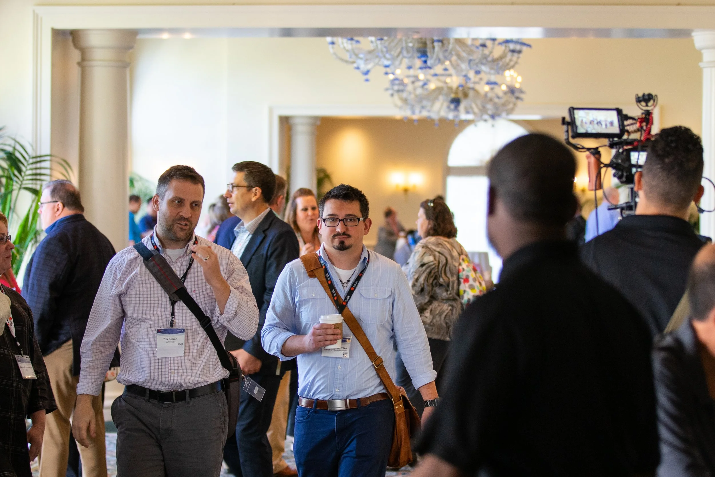 People gather at a convention or conference in an elegant room with chandeliers, columns, and natural light.