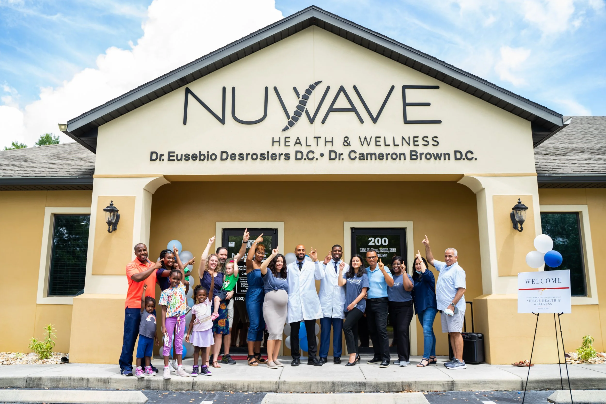 Group of people standing in front of a wellness center with a sign that reads 'NUWAVE HEALTH & WELLNESS.' The people are smiling and pointing upwards, celebrating the opening or event.