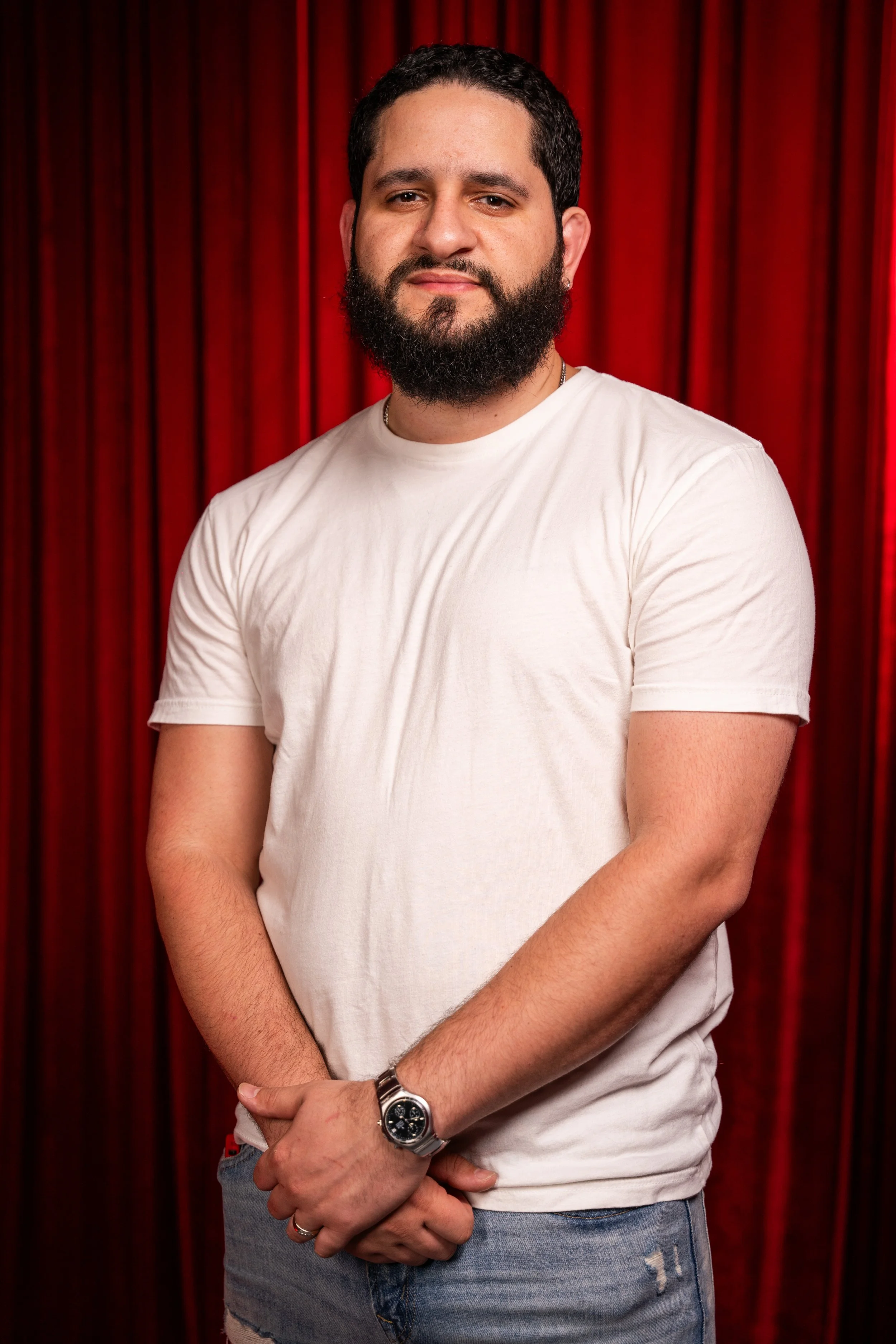 Portrait of a man with a beard, wearing a white T-shirt, standing in front of red curtains.