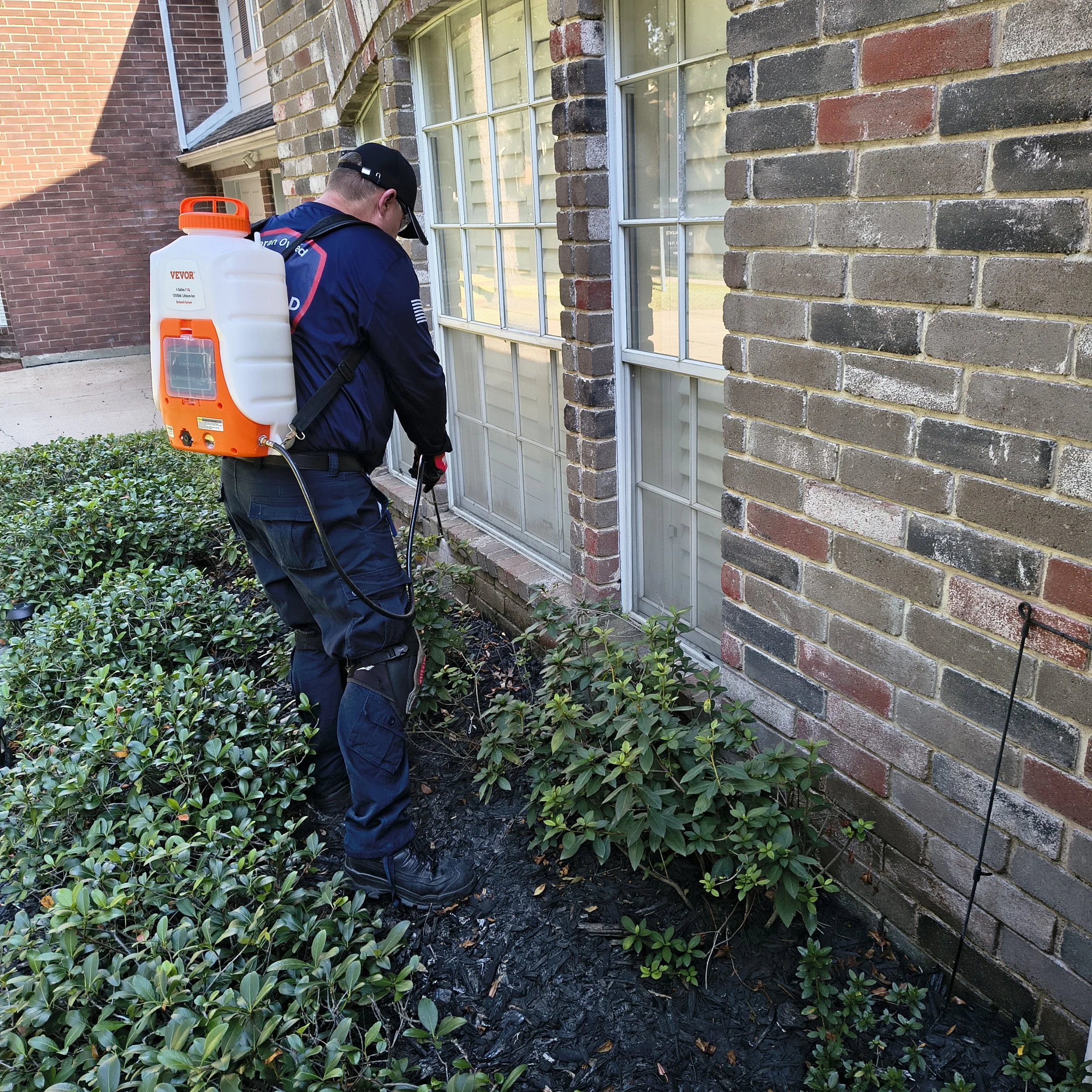 A person wearing a uniform and a backpack sprayer is spraying pesticide or herbicide on plants near a brick building.