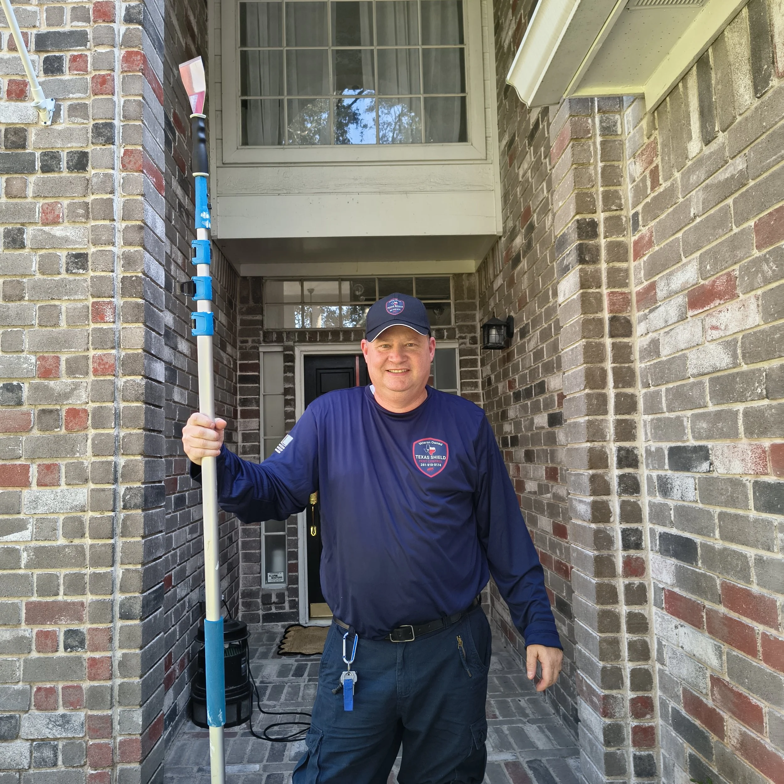 The owner of Texas Shield Pest Control in his uniform standing at the front door of a brick house, holding a cleaning or maintenance tool with a blue handle, smiling.
