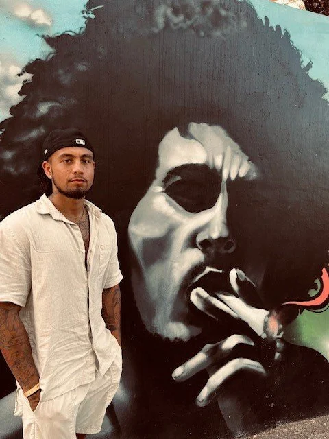 A man with a beard, wearing a light-colored shirt and a black cap, standing in front of a mural of Bob Marley.
