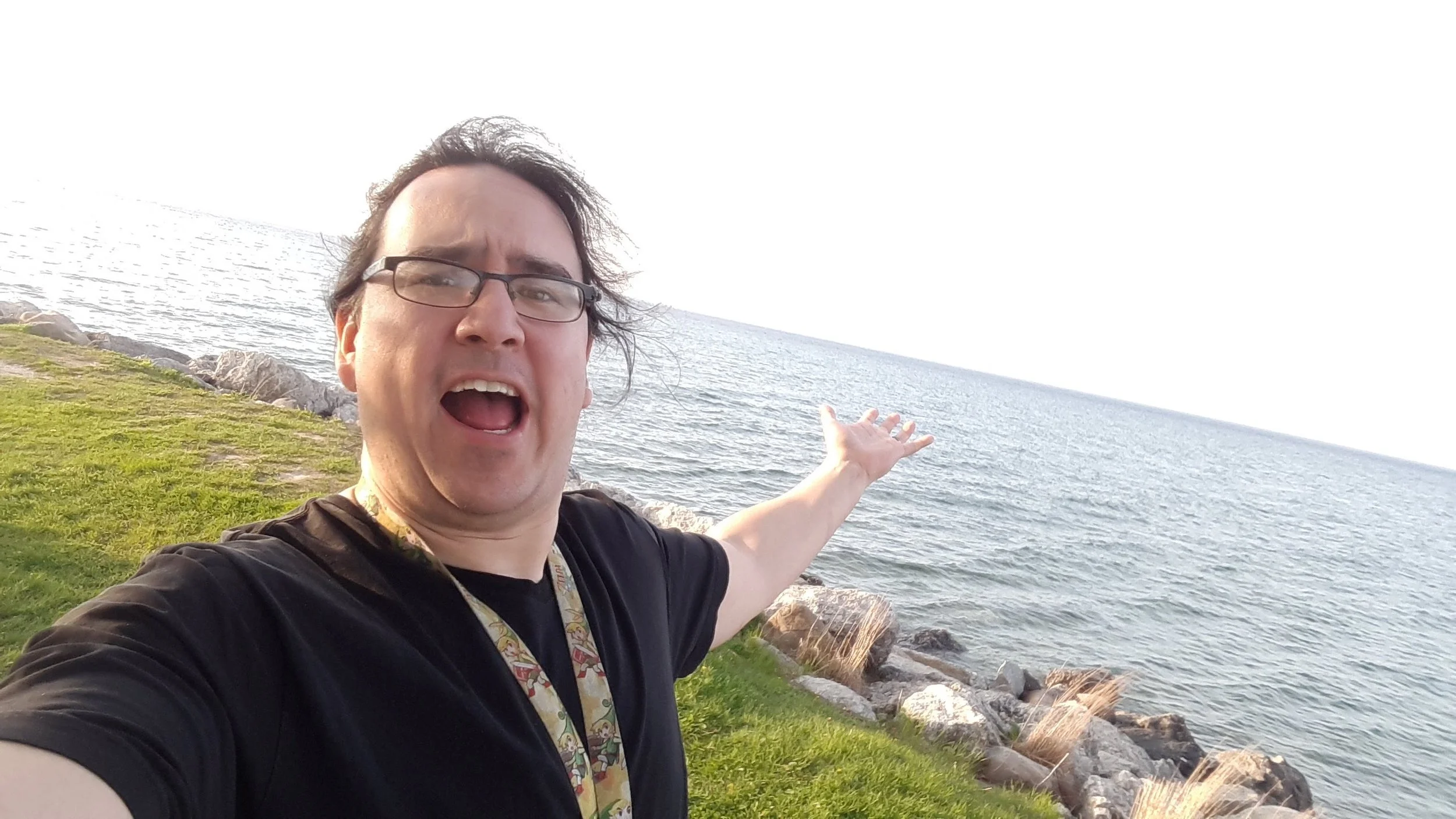 A man standing outdoors by the water with rocks and grass, extending his arm towards the ocean.