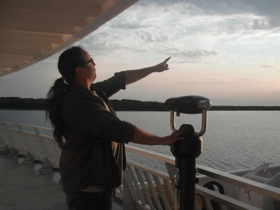 A man on a boat deck, pointing at the sky with a view of the lake in the background, during sunset.