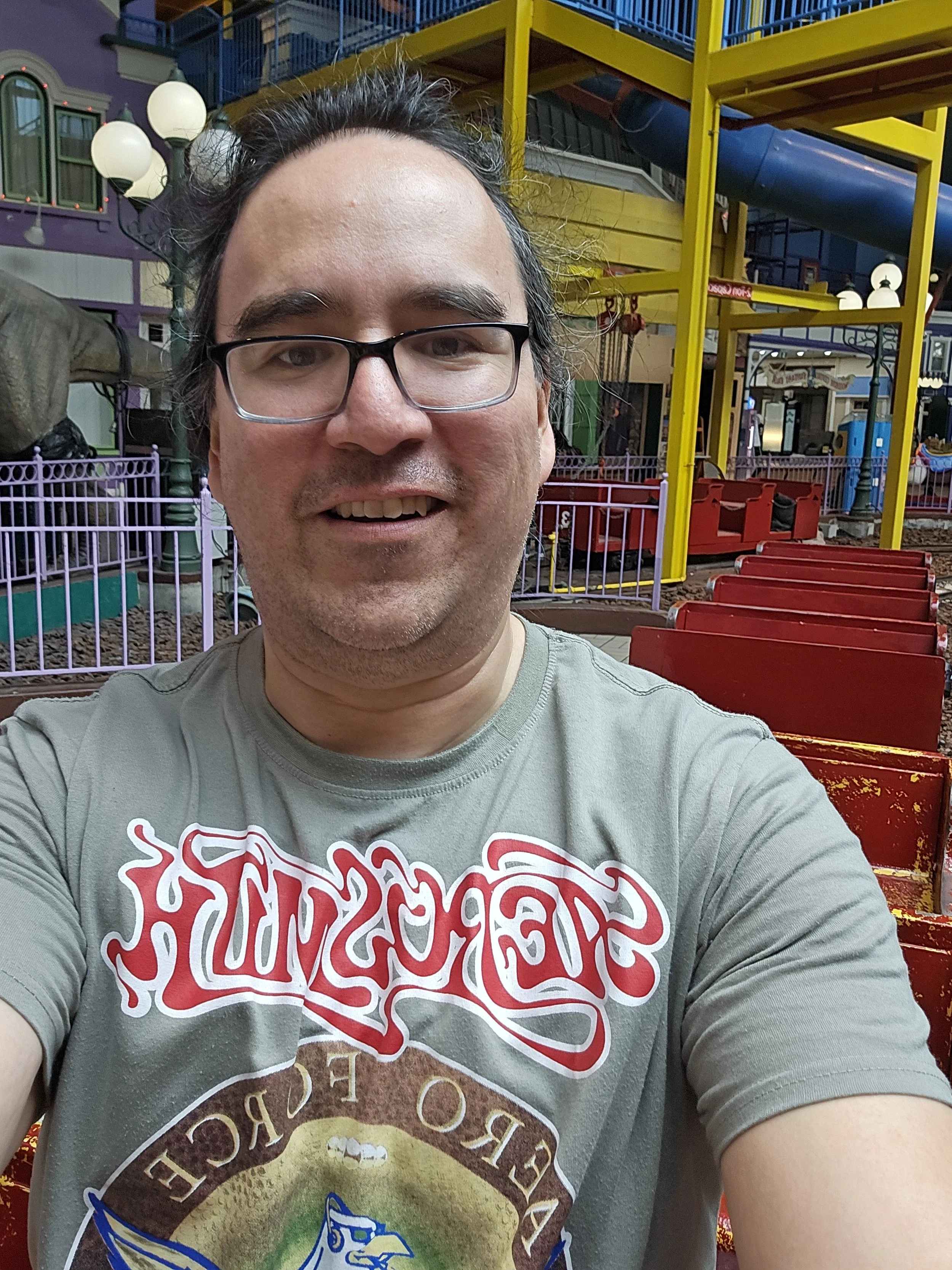 A man with glasses taking a selfie at an amusement park, with colorful rides and structures in the background.