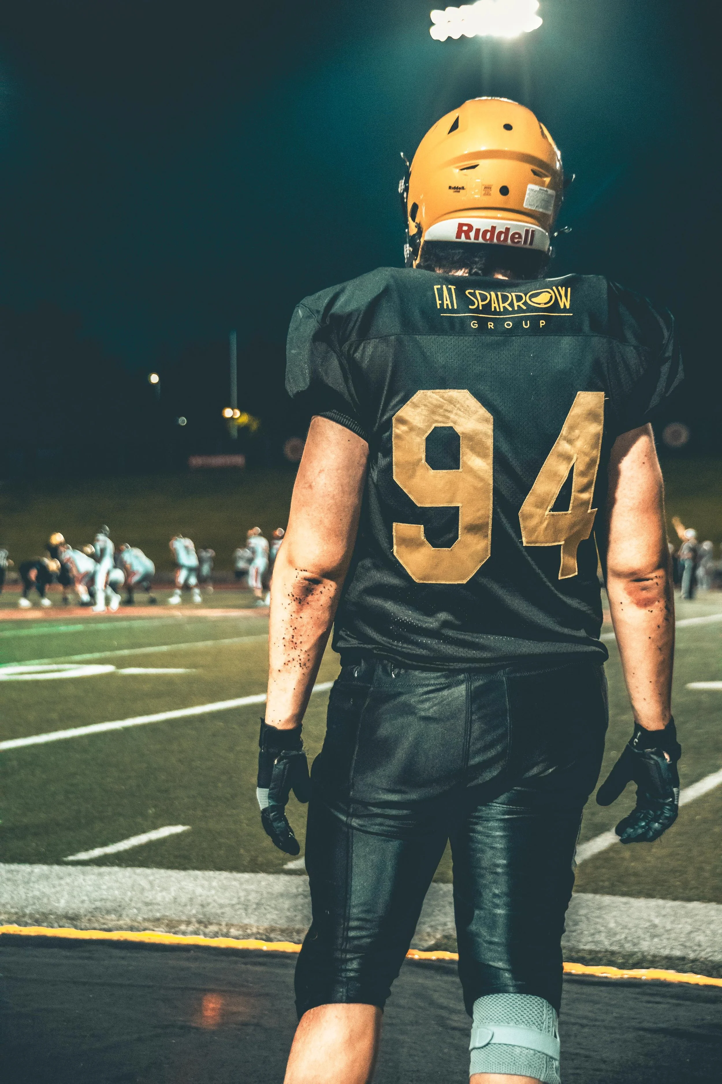 A football player wearing a black jersey with the number 94 and a yellow helmet, standing on the sideline during a game at night, with other players visible in the background.
