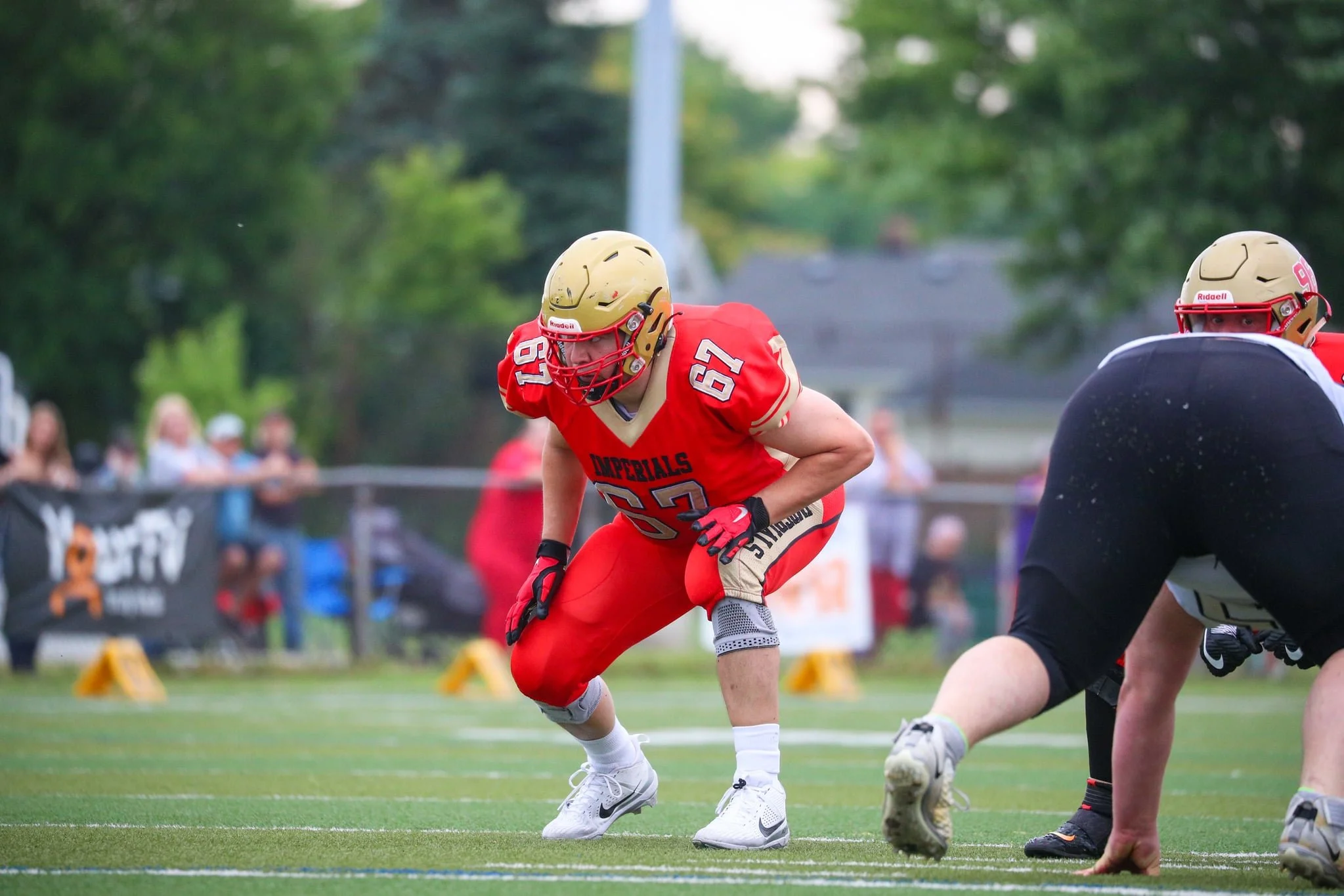 A football player in a red and gold uniform crouches on the field, preparing for the snap, with other players and spectators in the background.