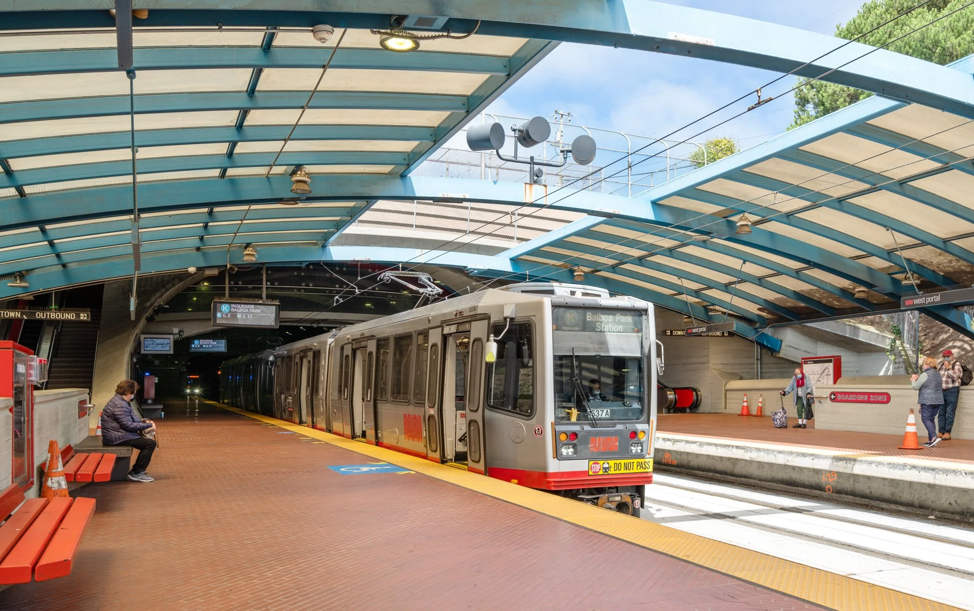 A light rail train at a station with a curved blue roof, yellow safety line, and people waiting on the platform. A man sits on a bench, and others stand with luggage near a sign that reads 'Boarding Zone'.