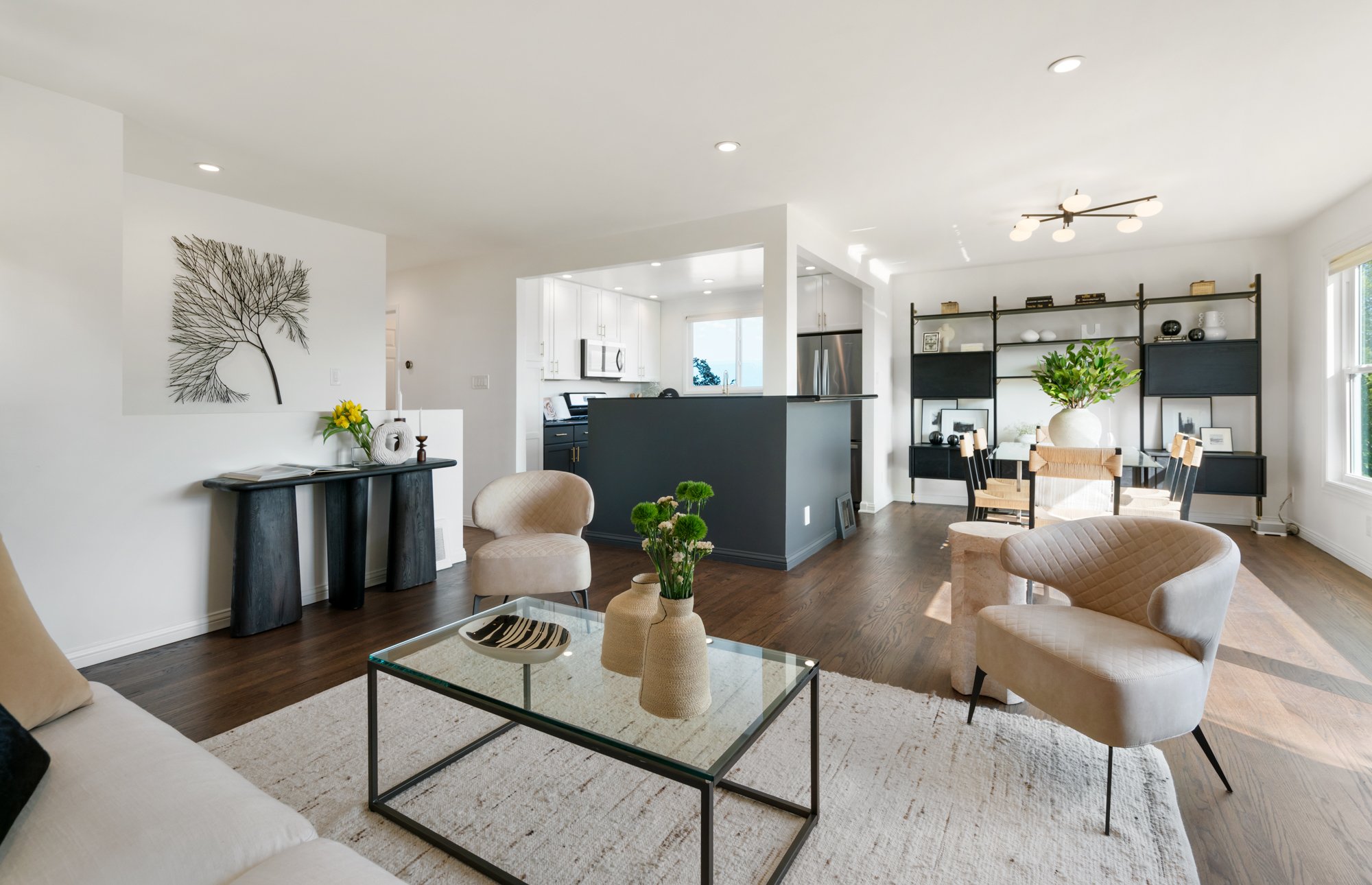 Living room with beige furniture, glass coffee table, and decorative vases, adjacent to a dining area with a black bookshelf and large windows