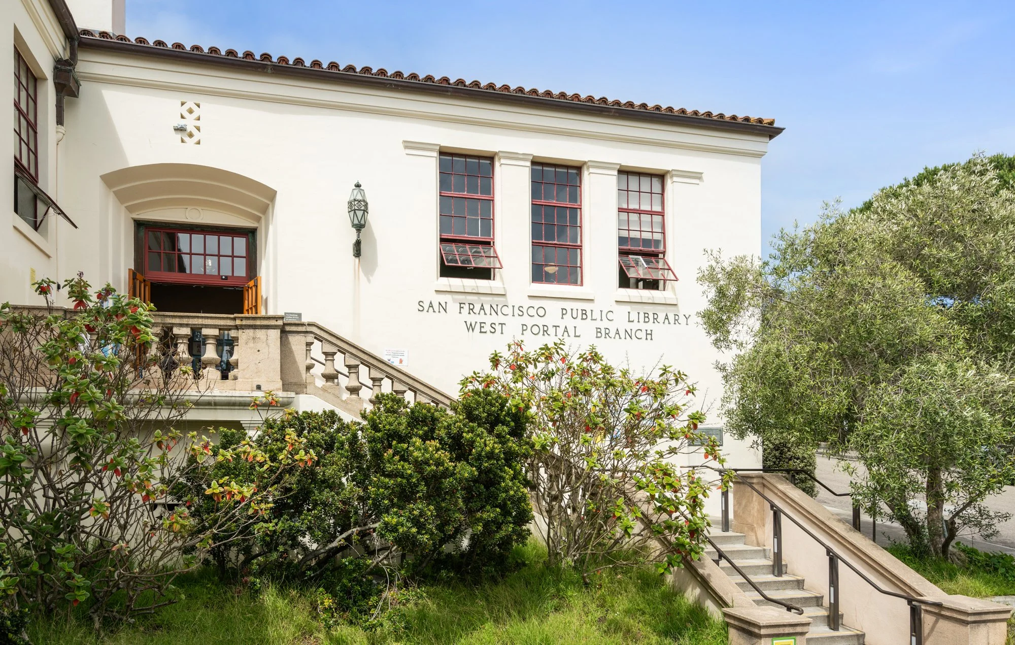 Exterior view of the San Francisco Public Library, West Portal Branch, showing the white building with three red-framed windows, a staircase, and surrounding greenery.