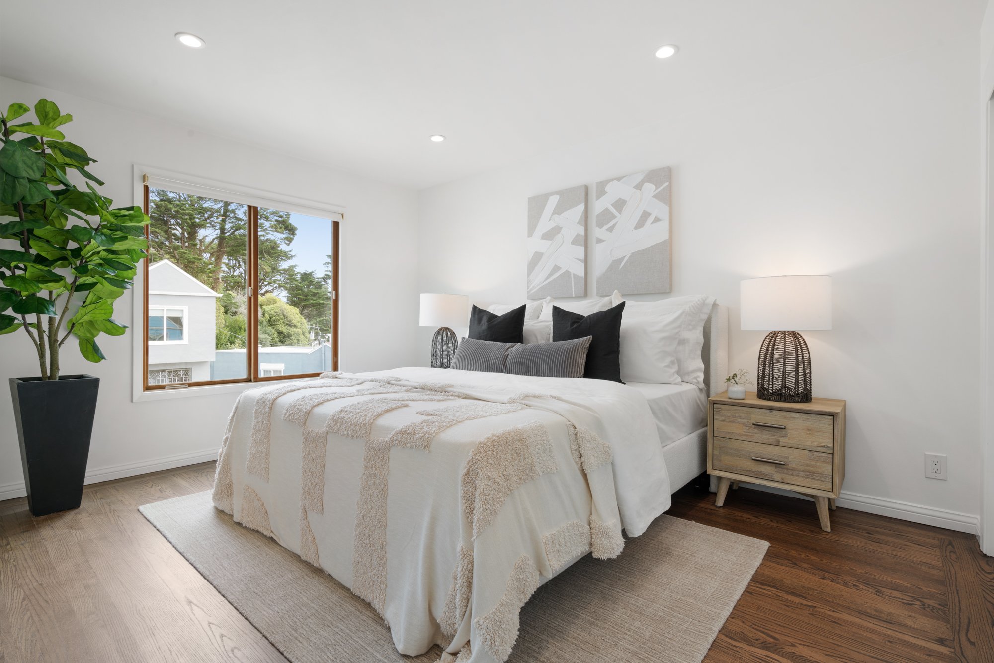 A bright bedroom with a large bed, white bedding, black and gray pillows, bedside table with a lamp, large window, wooden floor, and a potted plant.