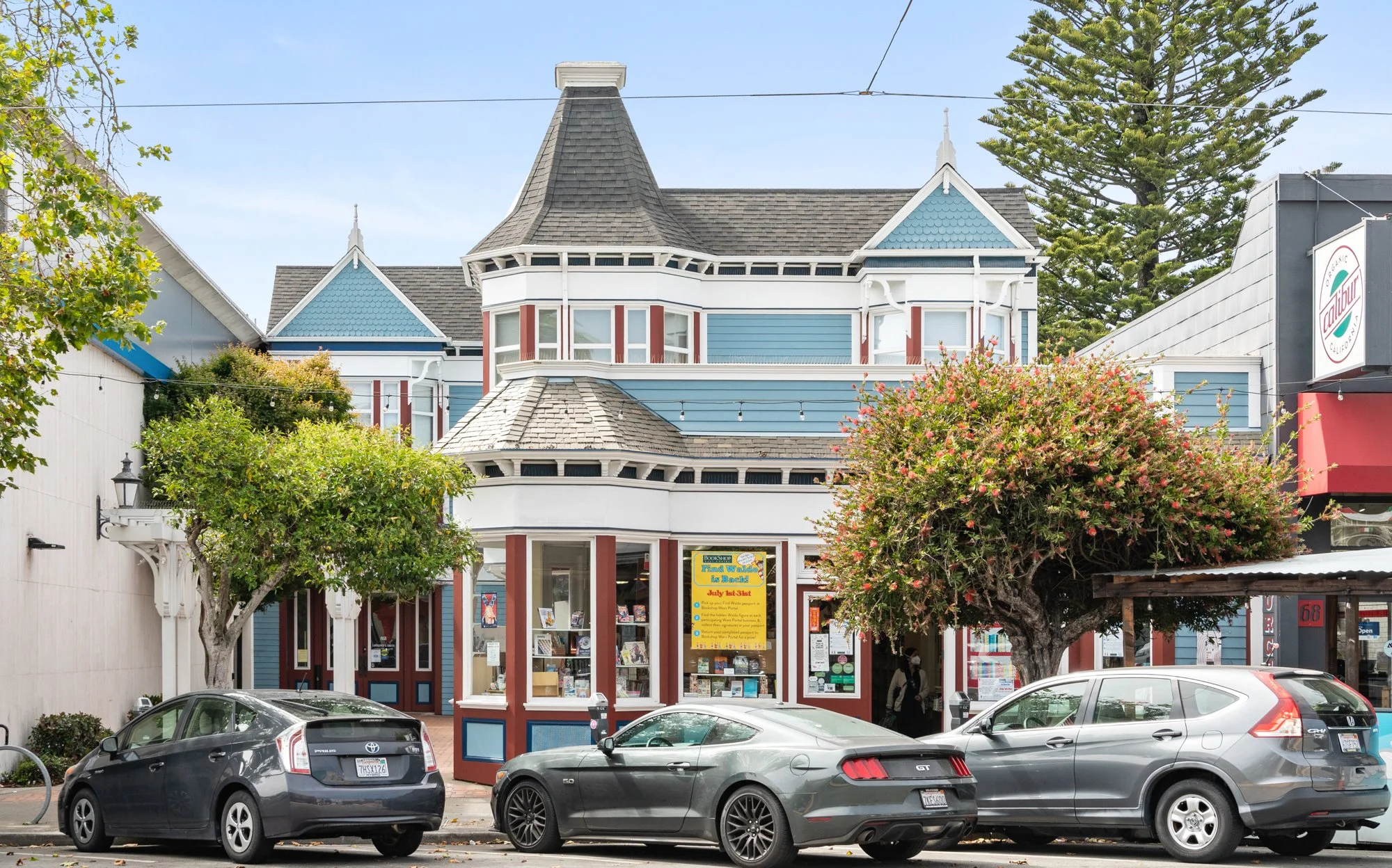 A Victorian-style building with blue and white exterior, bay windows, and a small corner shop on the ground floor, flanked by two trees and parked cars in front.