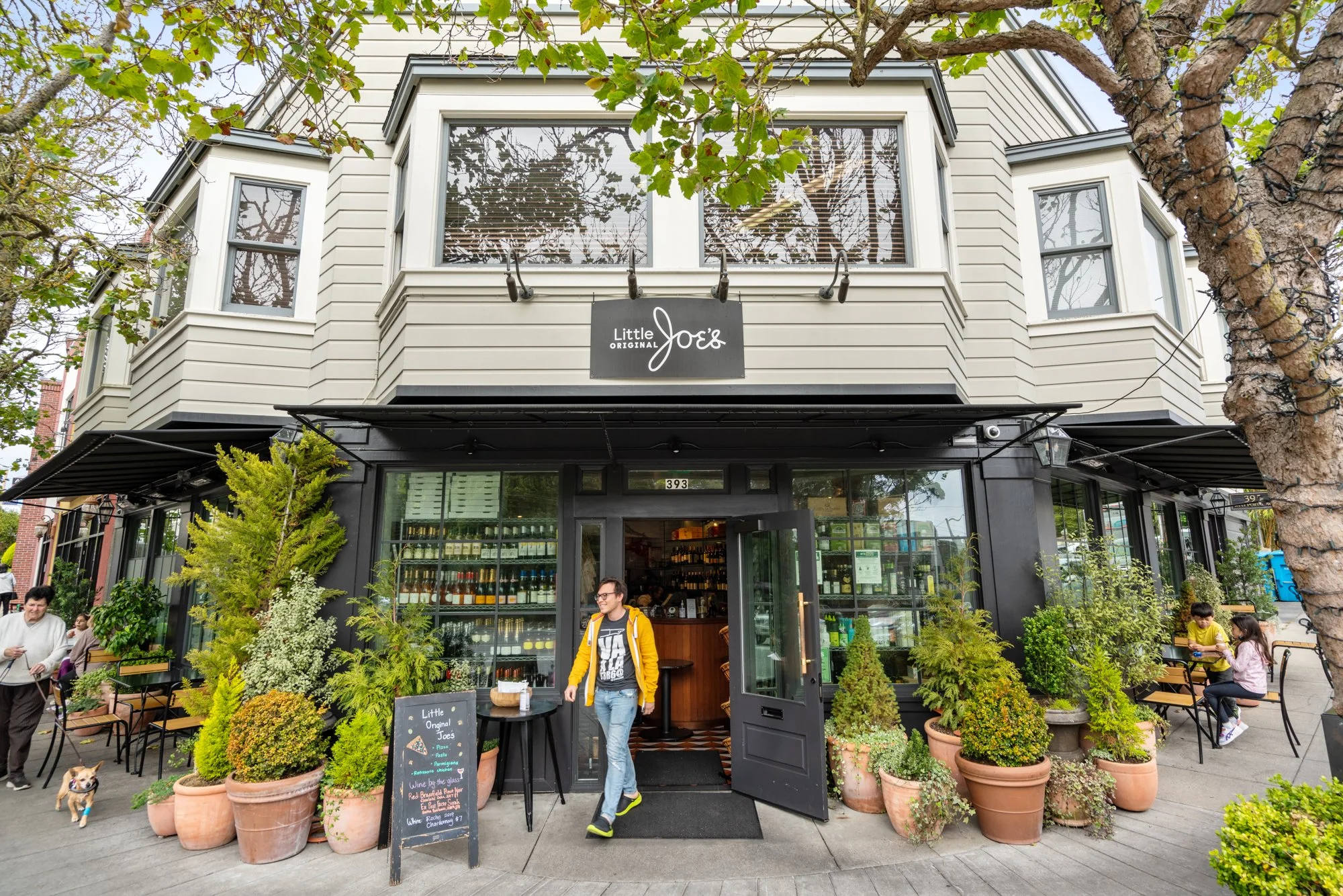 Exterior view of a two-story restaurant named Little Joe's with large windows and potted plants outside. A man in a yellow jacket is walking out of the restaurant, and a chalkboard sign is placed near the entrance. People are sitting and walking arou