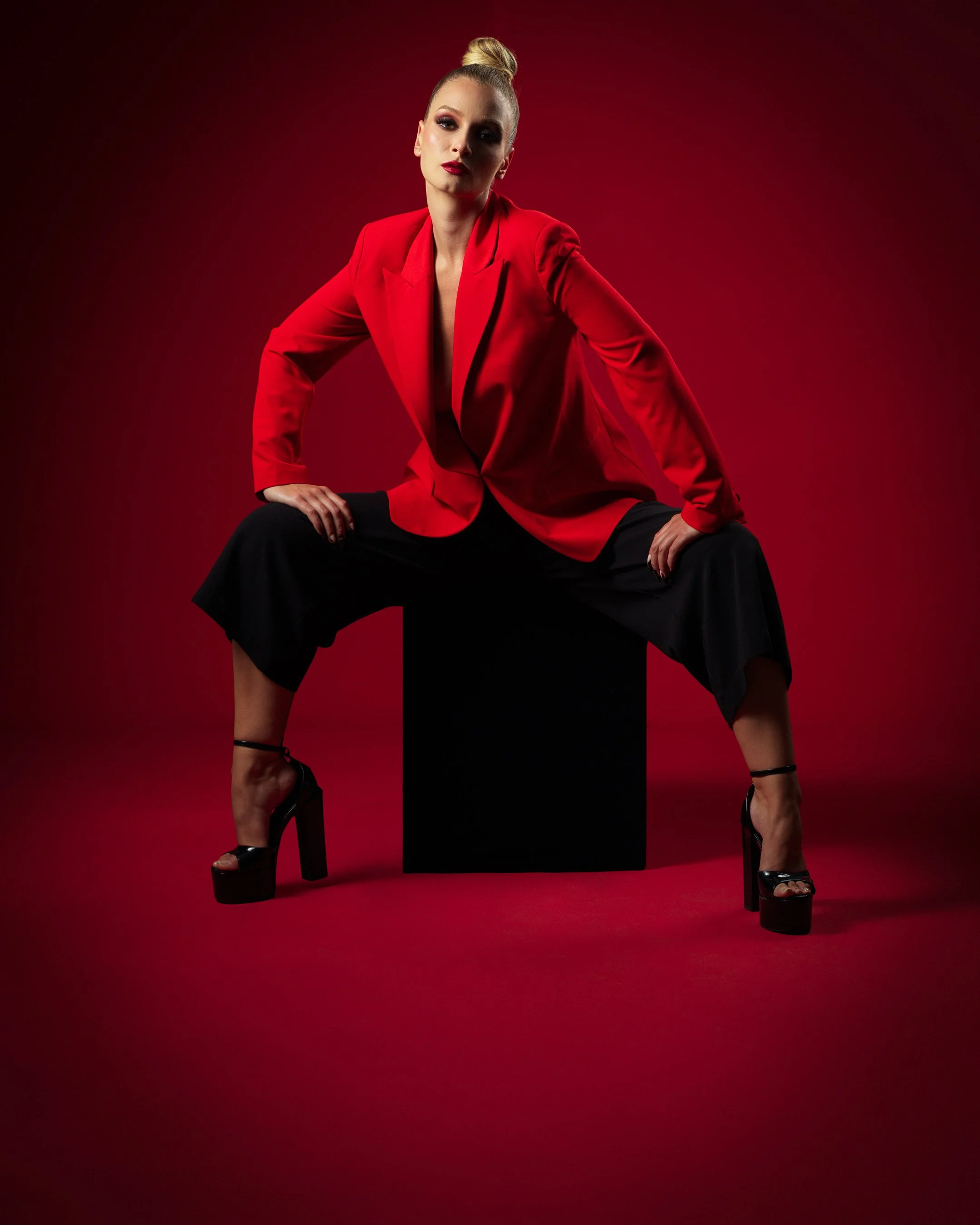 Crimson Authority — fashion editorial by Peter Lovit. Model wearing a red tailored blazer and black trousers, seated against a dramatic crimson studio background.