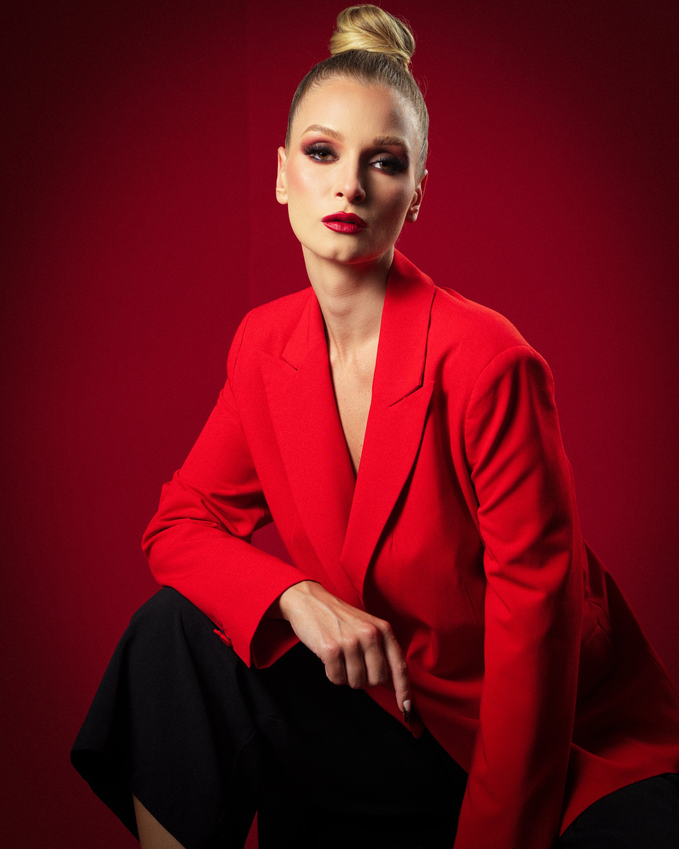 Crimson Authority — fashion editorial by Peter Lovit. Model wearing a red tailored blazer and black trousers, seated against a dramatic crimson studio background.