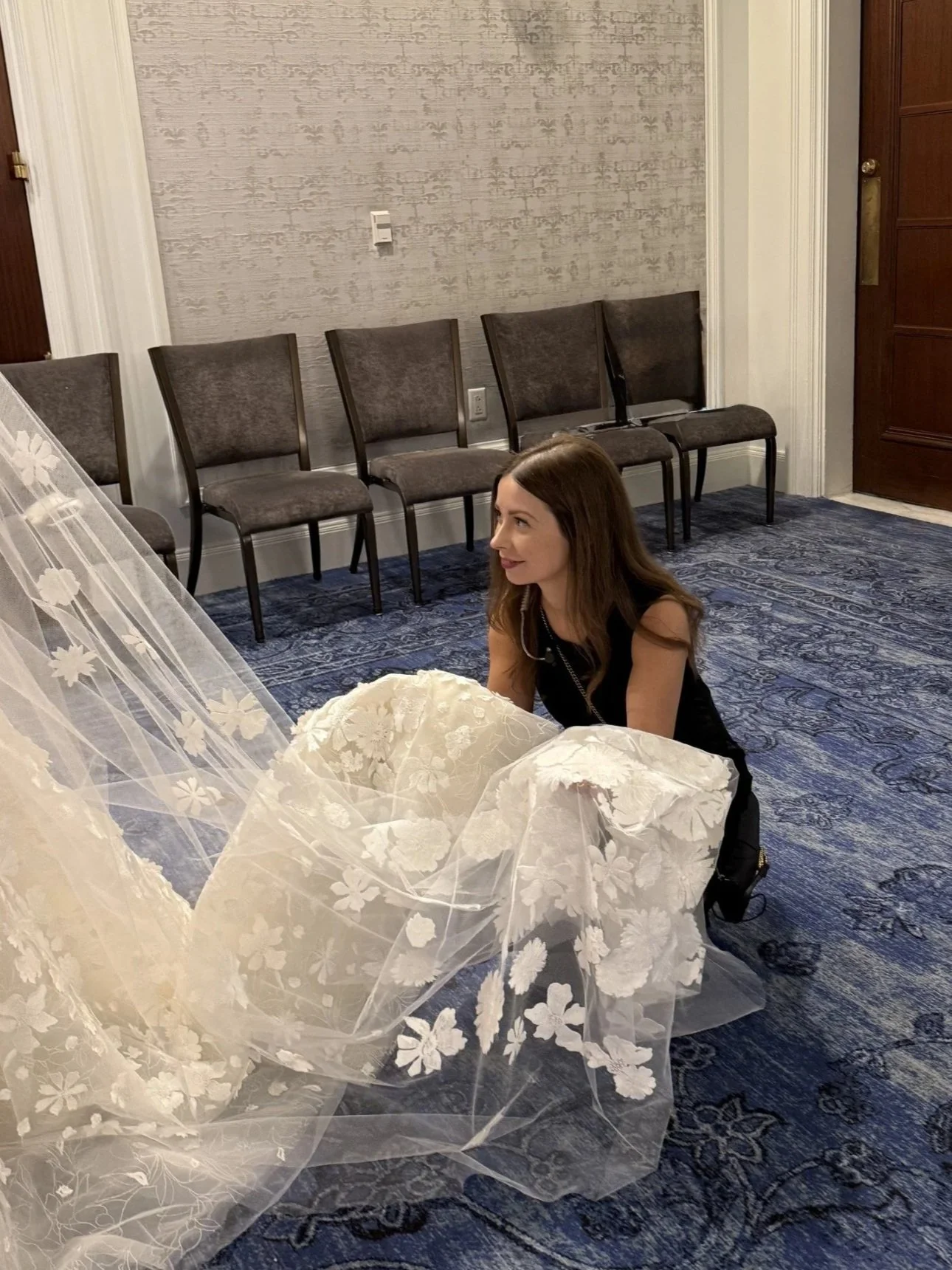 A woman with long brown hair, wearing a black dress, smiling and kneeling on the floor next to a white, floral-patterned wedding veil.