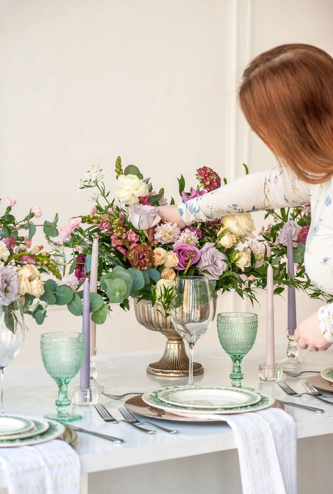 Rebecca Buford arranging a floral centerpiece on a decorated dining table with green and purple glassware, white plates, silverware, pink candles, and a white table runner.