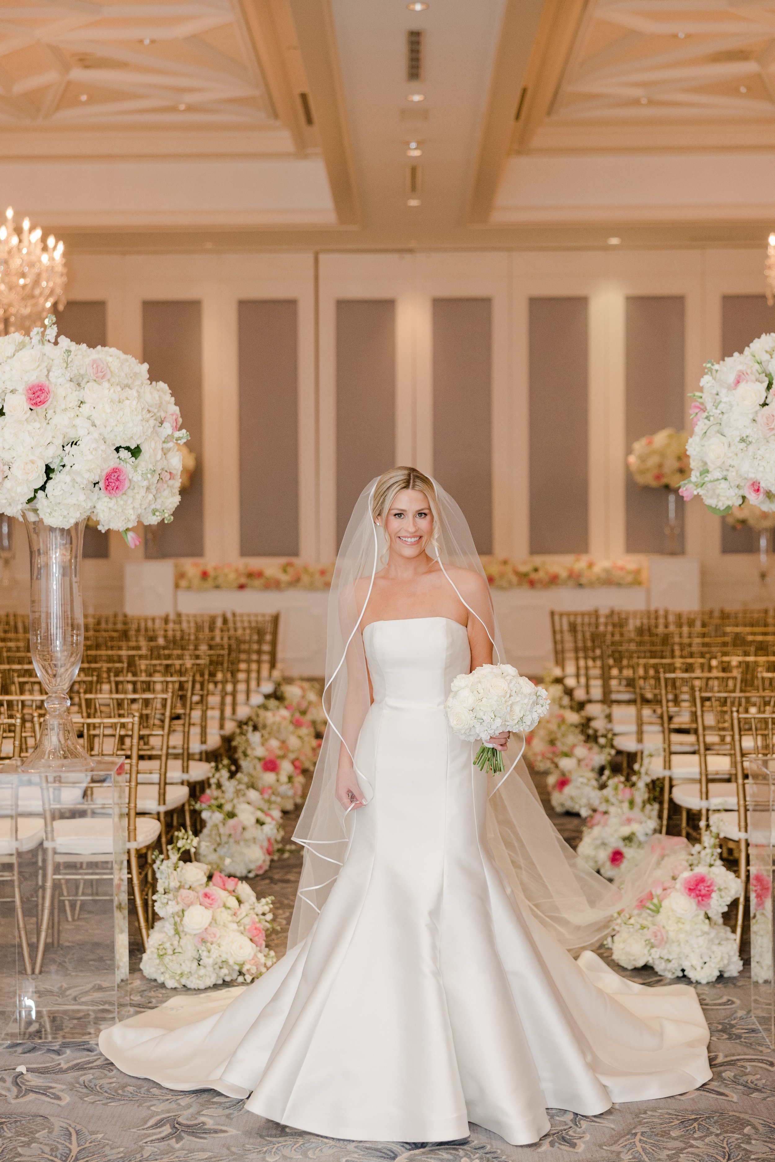 A bride in a white wedding gown holding a bouquet stands in a decorated wedding venue.