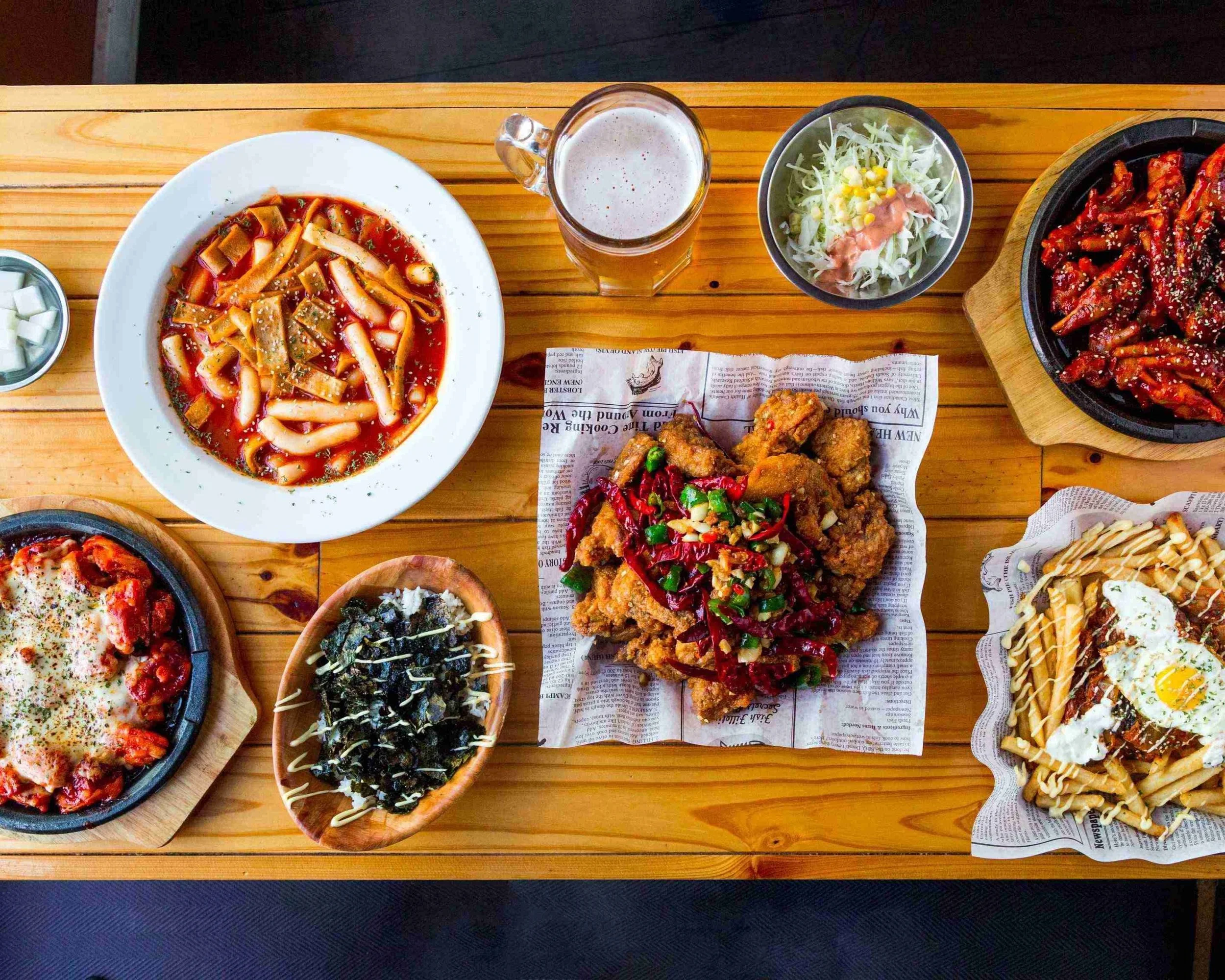 Assorted Korean dishes on a wooden table, including tteokbokki, fried chicken, seafood pancake, pasta, salad, and beer.