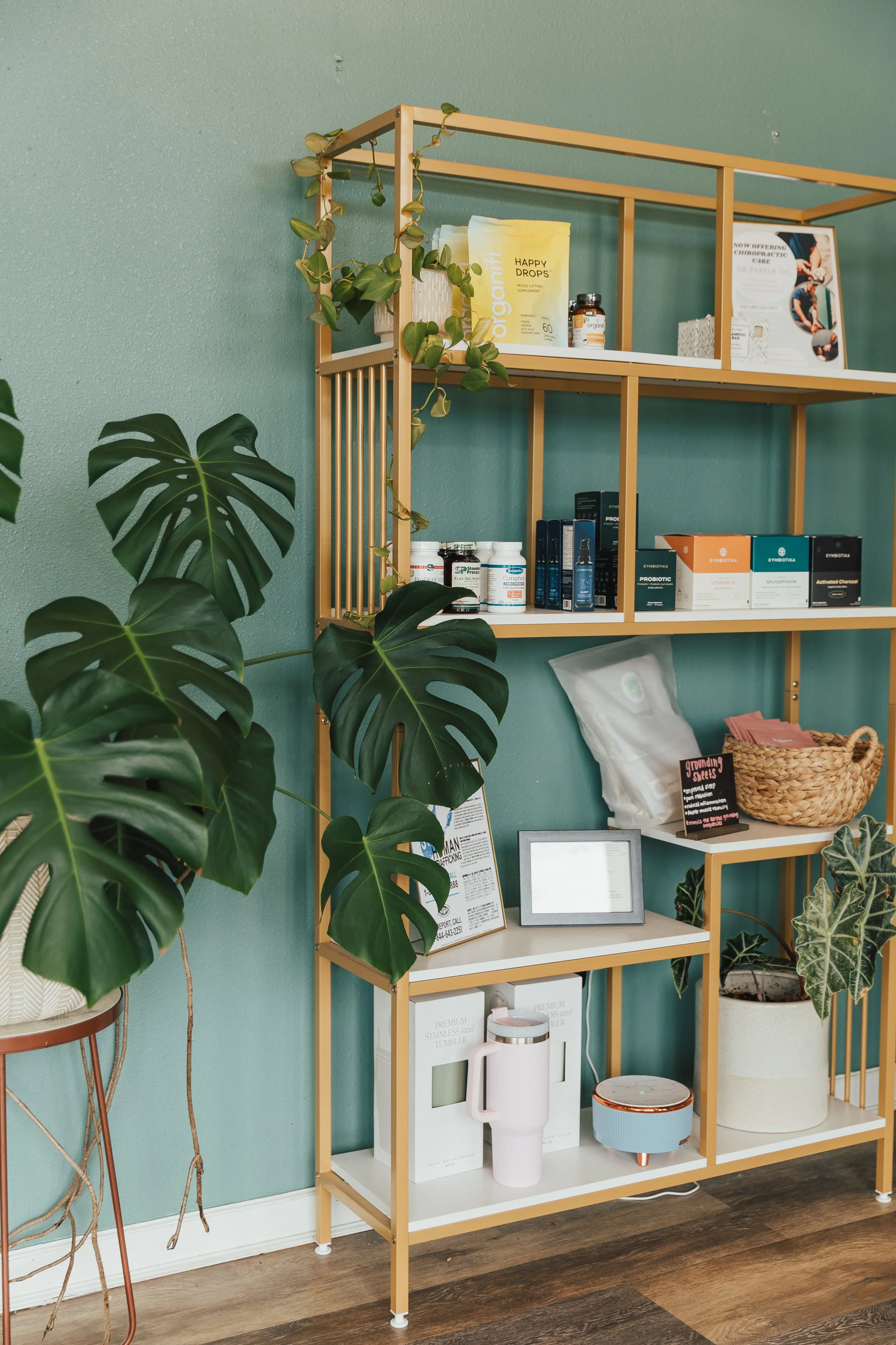 A wooden shelving unit with various health and wellness products, a framed tablet, a basket with pink items, and green plants against a teal wall.