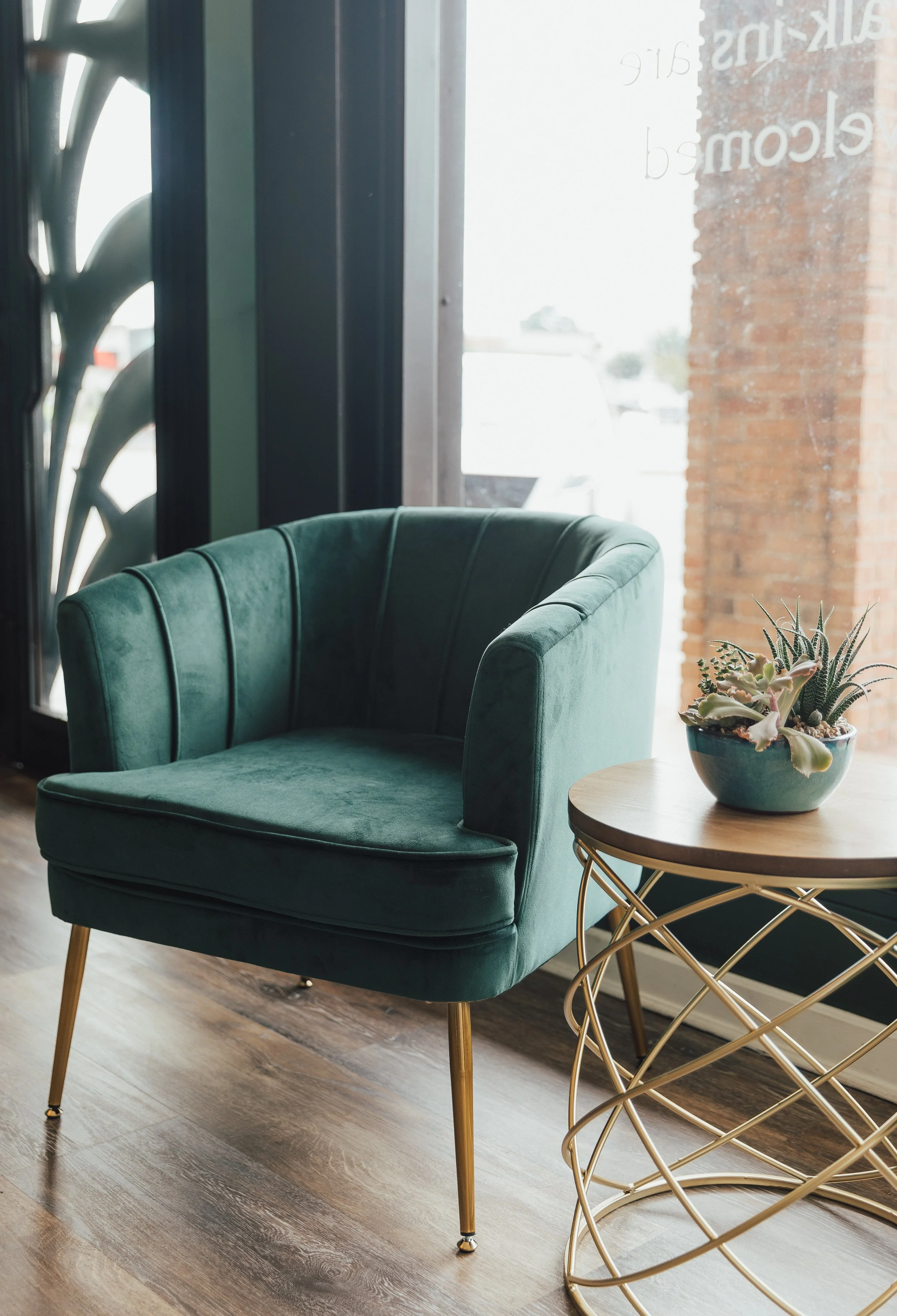 Green velvet armchair with gold legs next to a round wooden table holding a succulent plant in a blue bowl, against a glass window with visible outdoor scene.