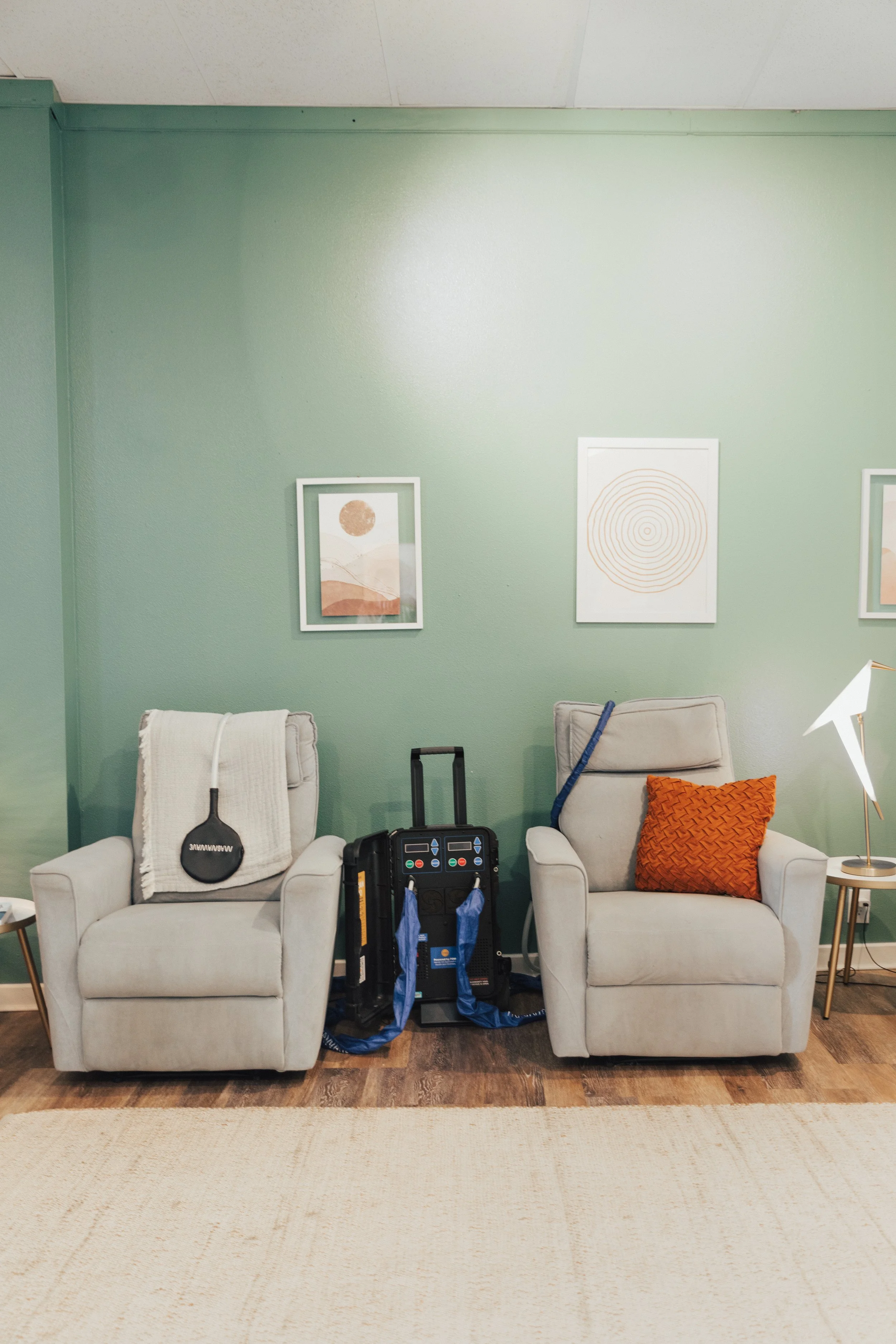 Two beige armchairs with a small side table and decorative pillows, placed against a pastel green wall with abstract art, and a targeted pulsed electromagnetic field therapy device in between.