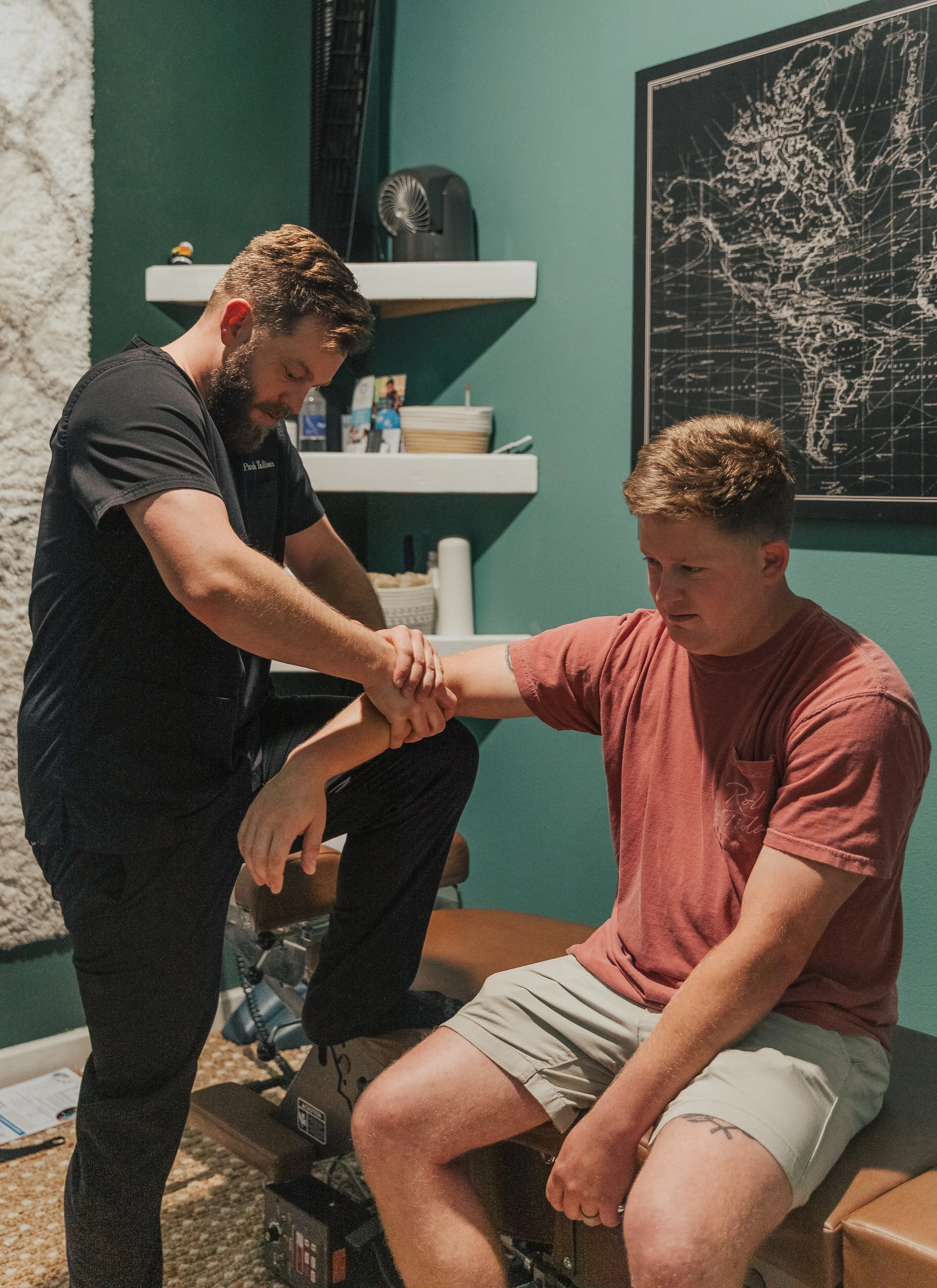 A chiropractor adjusting a young man’s arm while he sits on an examination table in a room with teal walls, a chalkboard map of South America, and shelves with medical supplies.