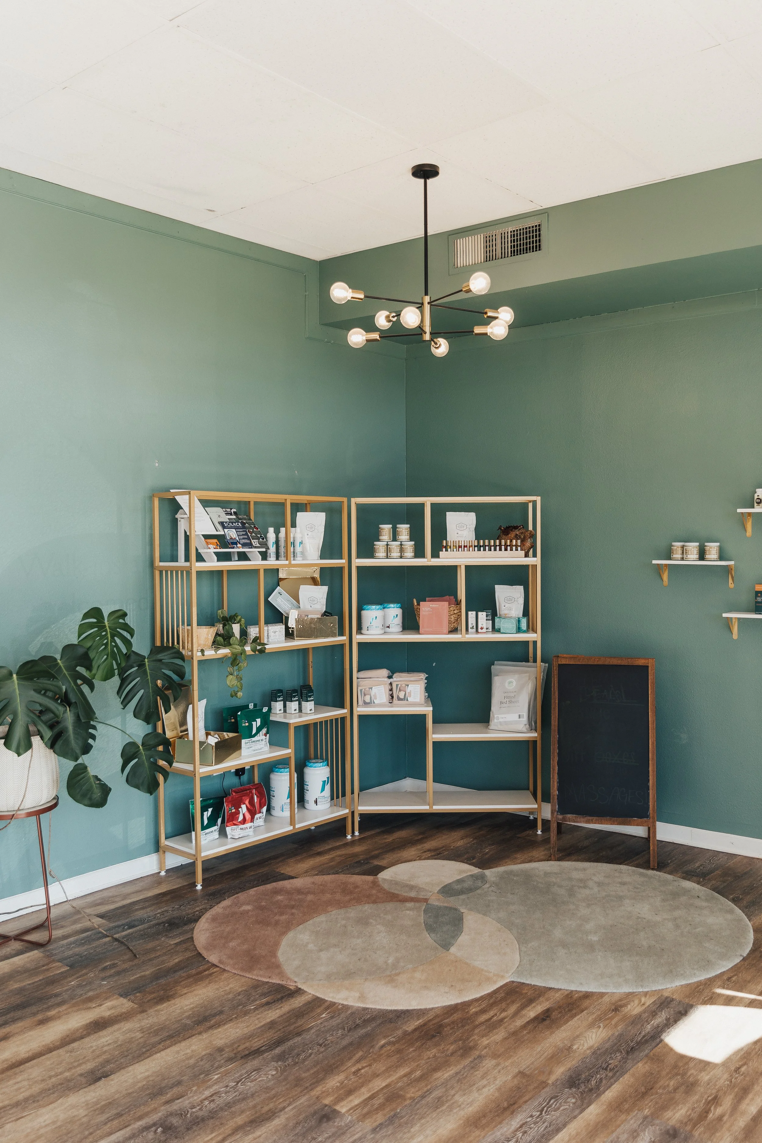 Interior of a wellness or retail store with green walls, wooden shelves displaying health products, a large green potted plant, a patterned rug, and a modern chandelier lighting fixture.