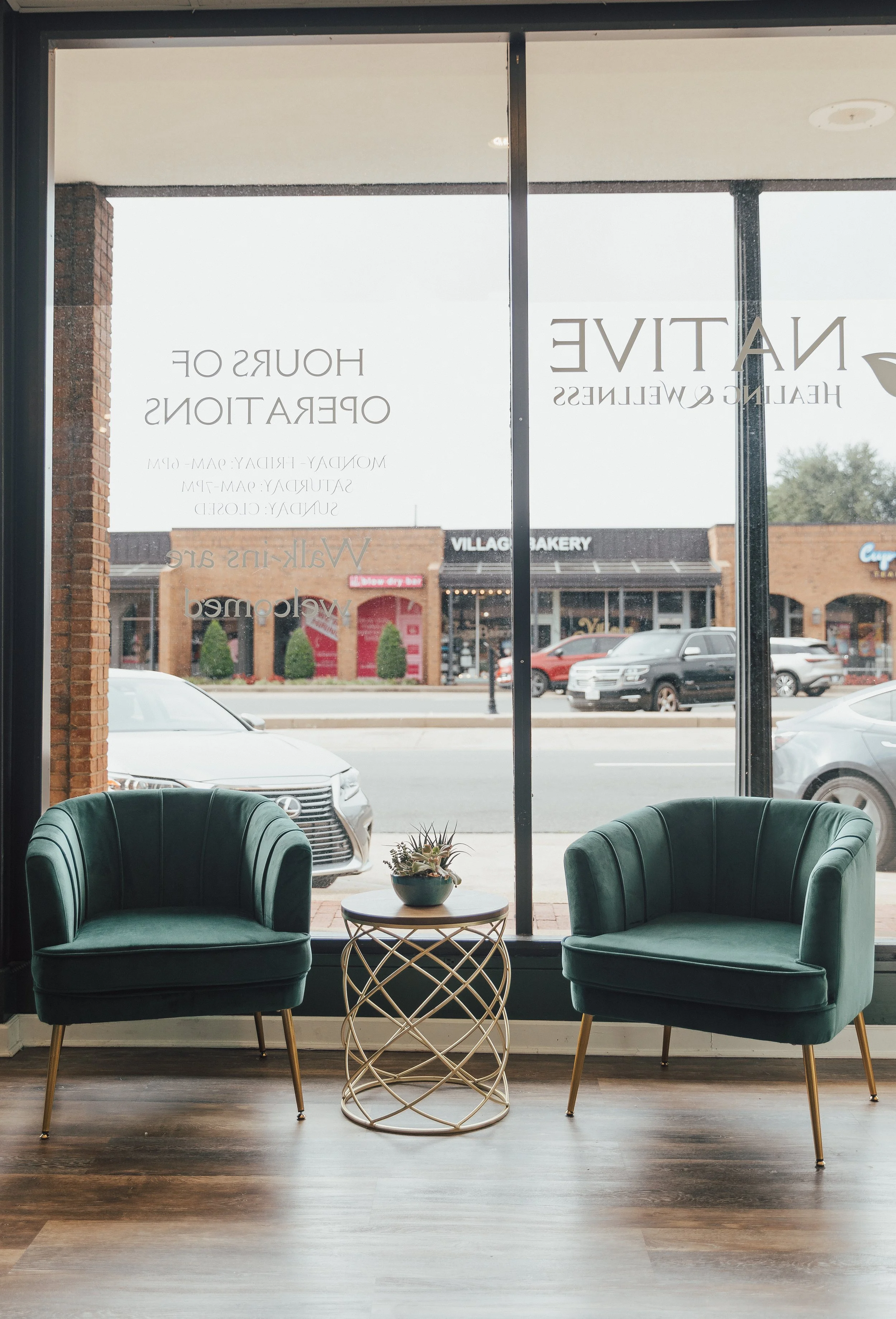 Two green upholstered armchairs with gold legs are positioned in front of large glass windows. Between the chairs is a small, round, gold wire-frame table with a small potted plant on top. Outside the window, parked cars and a row of storefronts are