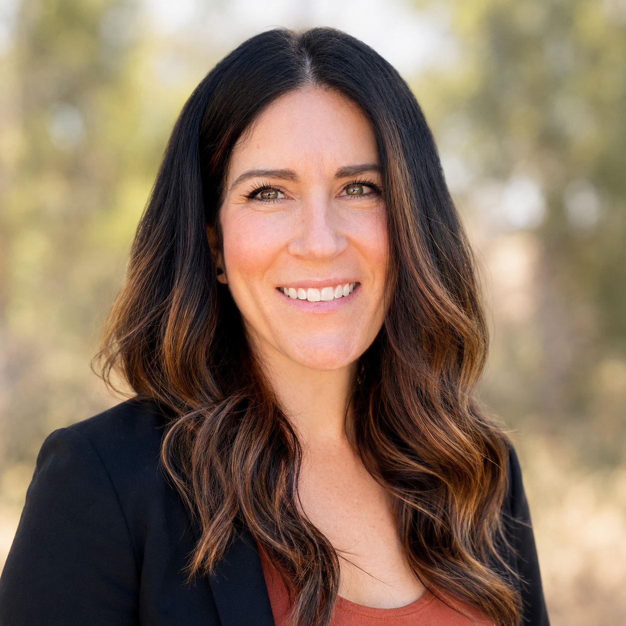 A woman with long, wavy dark brown hair and hazel eyes smiling outdoors in a natural setting.