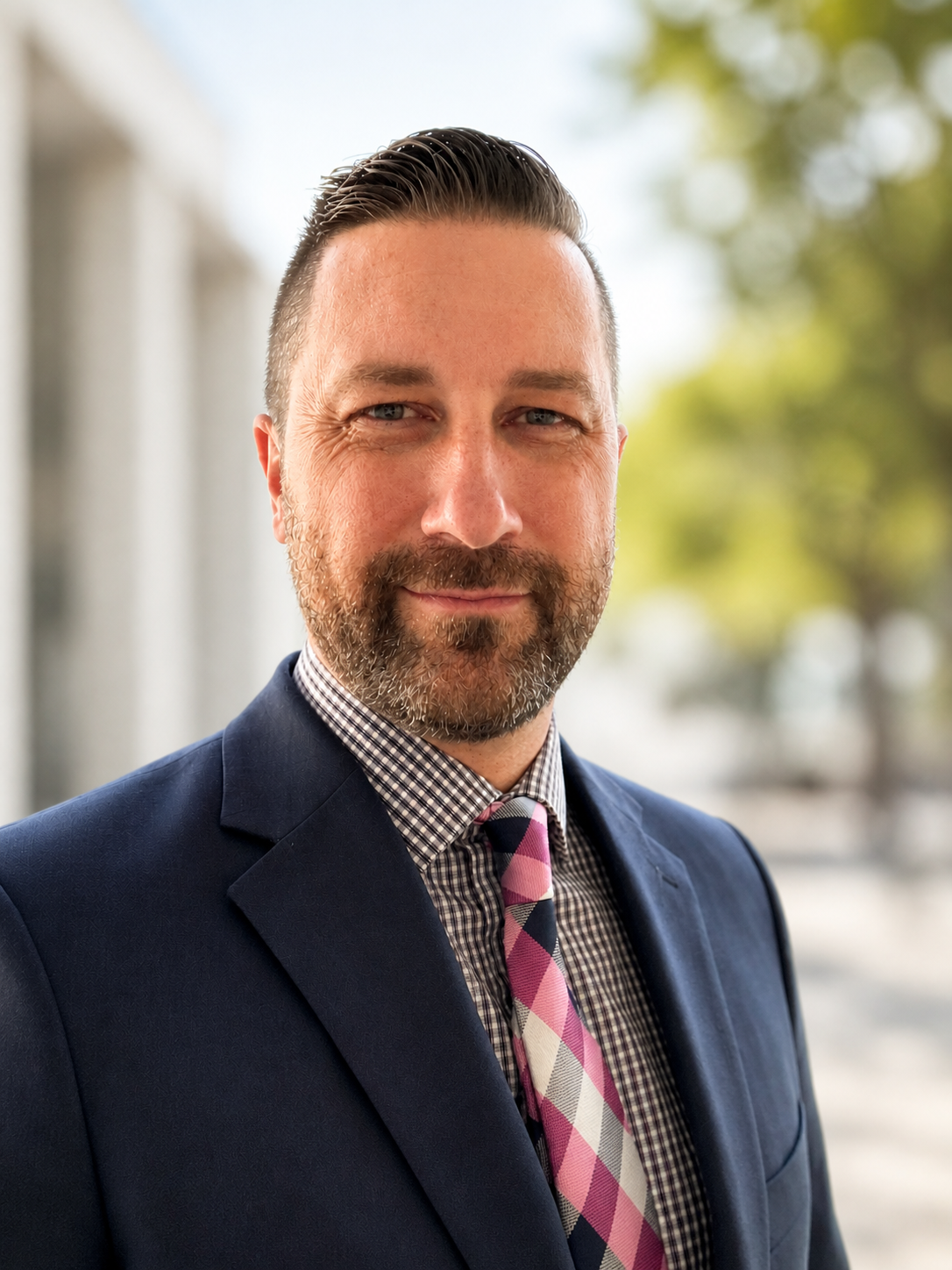 A portrait of a middle-aged man with a beard, wearing a dark suit, a checked shirt, and a pink plaid tie, standing outdoors with blurred trees in the background.