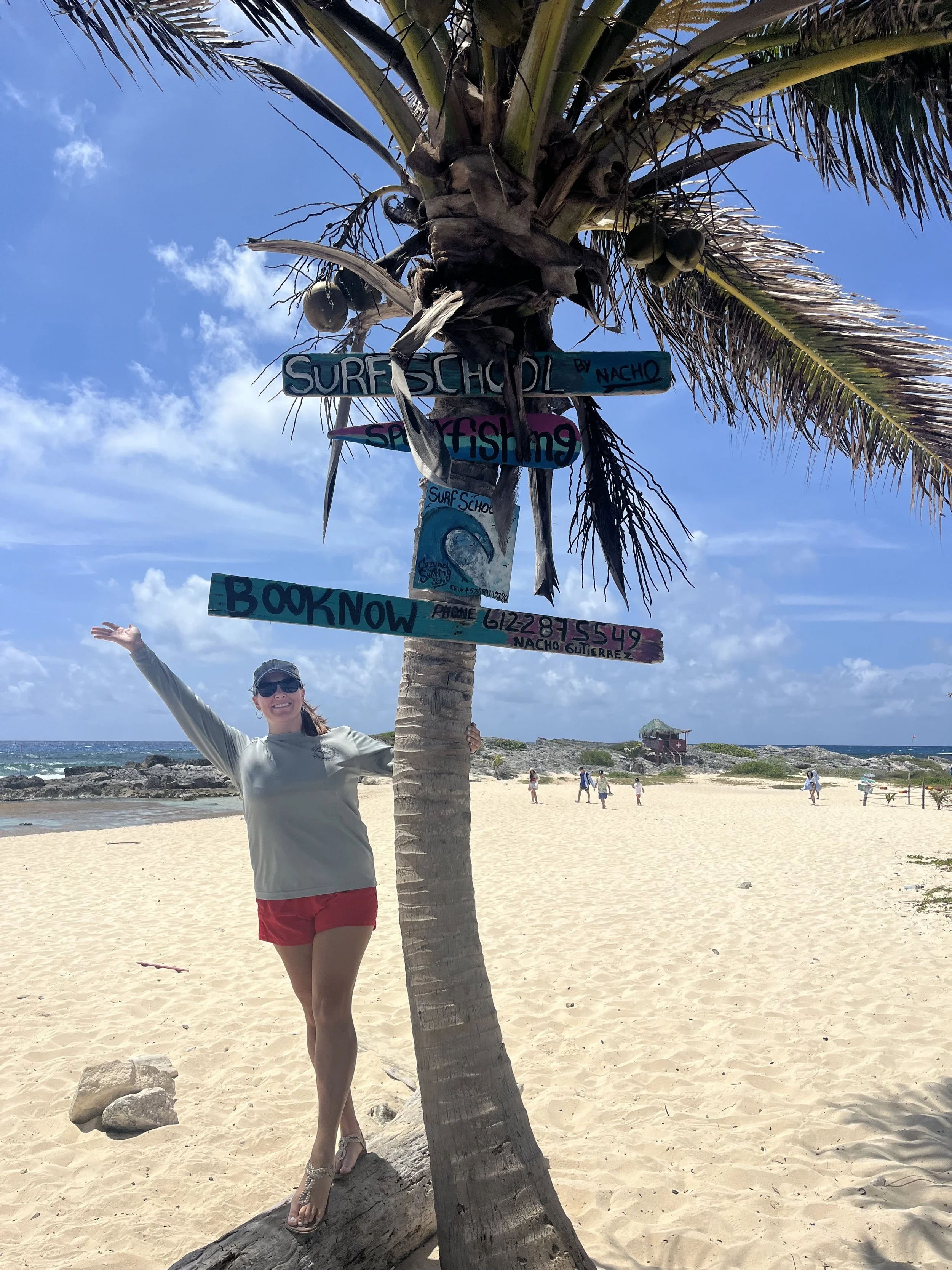 Woman in sunglasses and red shorts standing next to a palm tree with colorful signs promoting surf school, snorkeling, and booking information on a sandy beach under a partly cloudy sky.