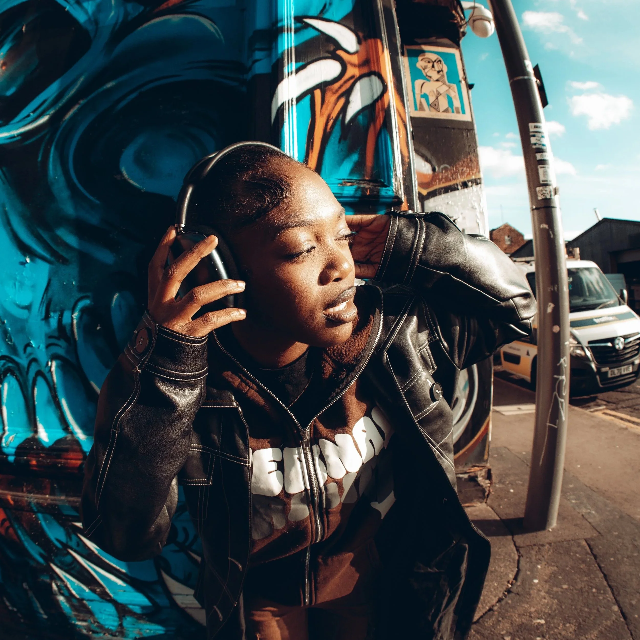A woman with closed eyes is listening to music with headphones while leaning against a graffiti-covered wall on a city street.