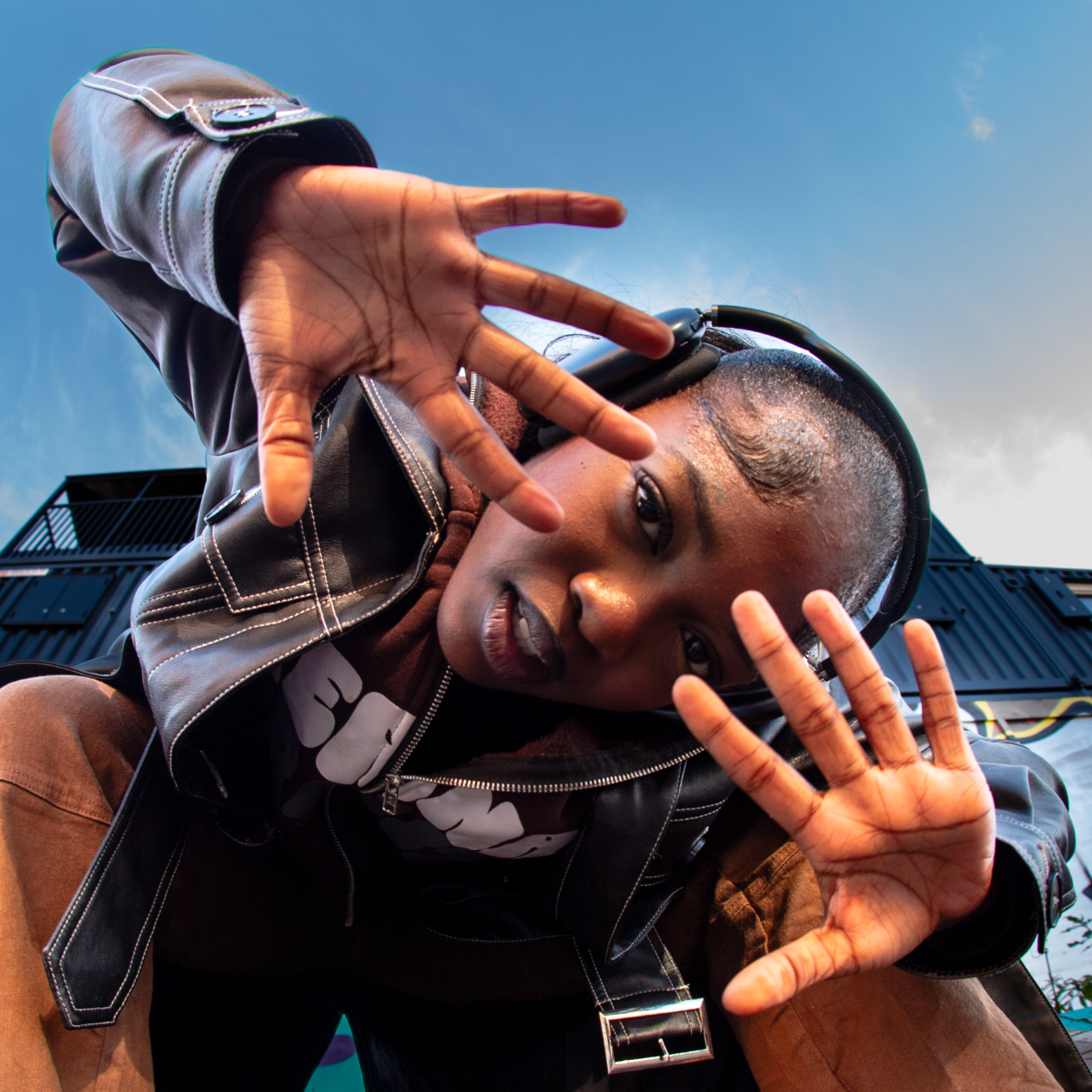 A woman with a shaved head wearing headphones, a leather jacket, and a brown shirt, squatting outdoors with a blue sky and a metal building in the background, reaching towards the camera with both hands.