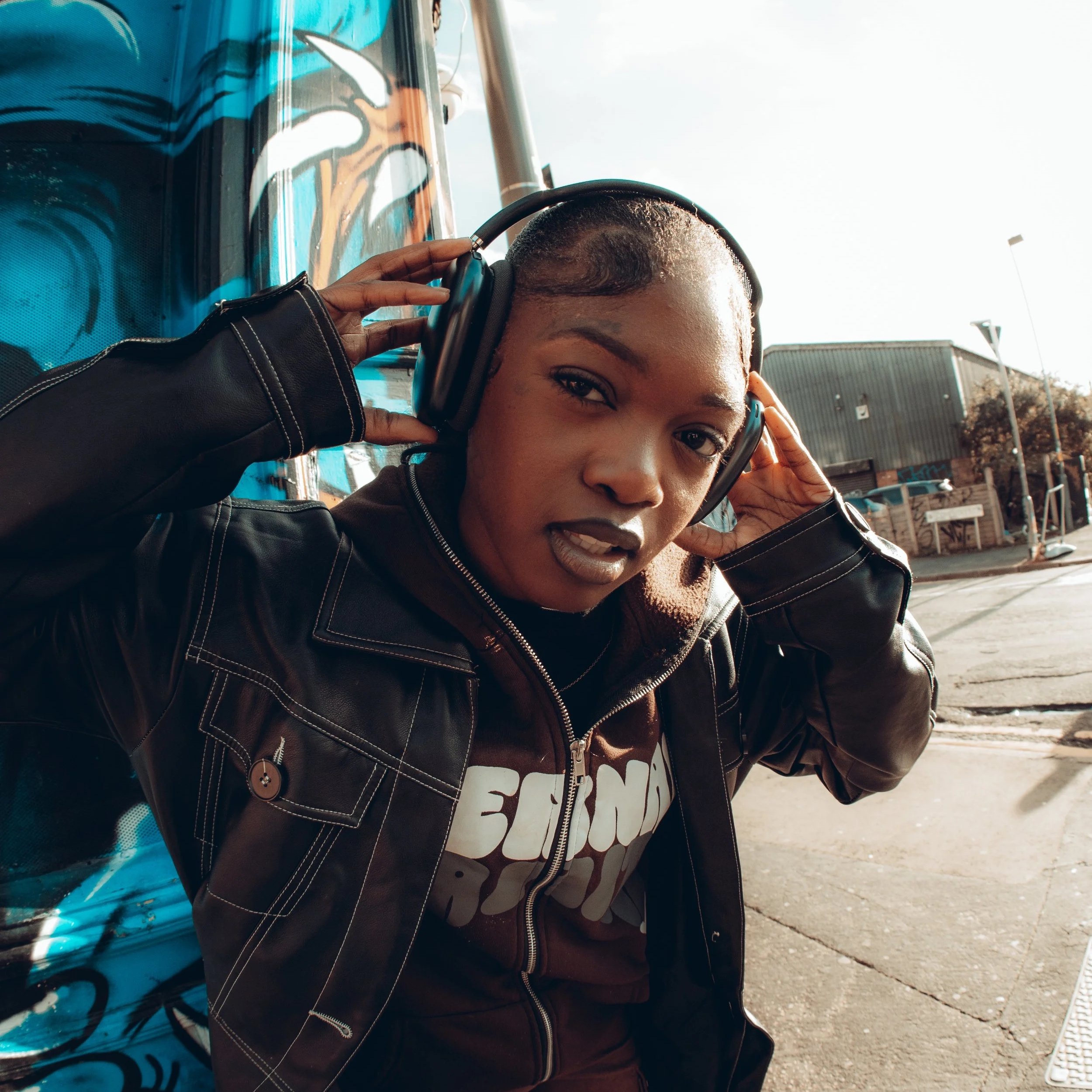 A young woman with short hair wearing a black leather jacket and headphones, standing on the street near a colorful mural, holding the headphones with both hands, looking at the camera.