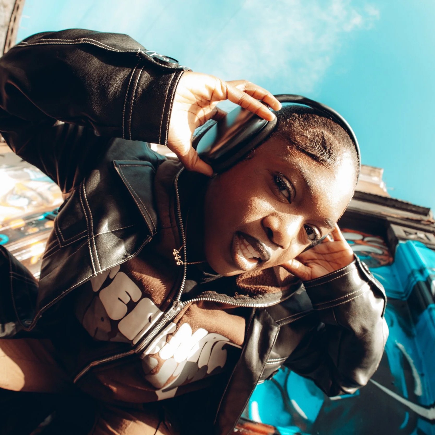 Young woman listening to music with headphones, wearing a black leather jacket, posing indoors with a blue wall and stacked items in the background.