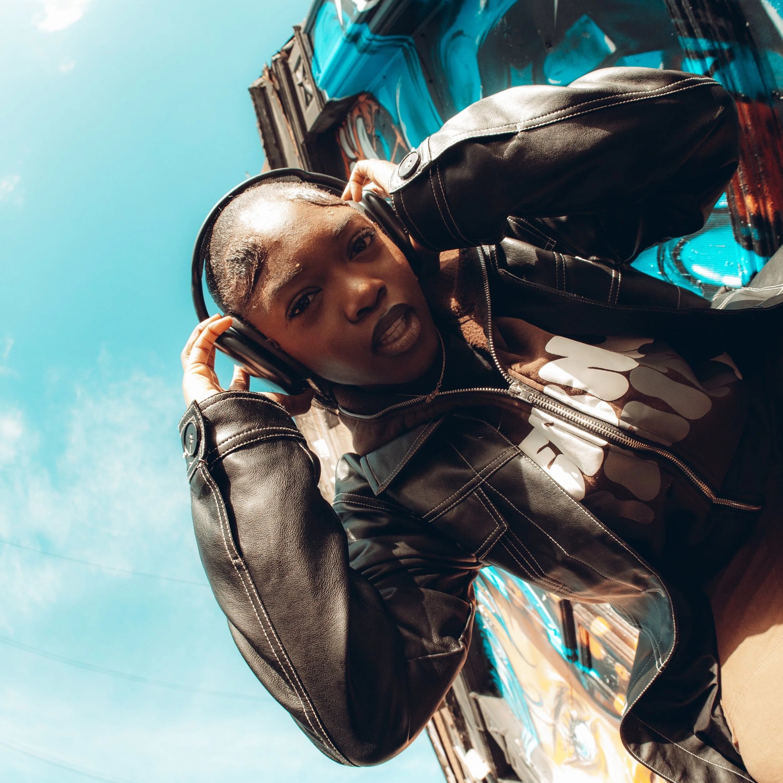 A young woman with braided hair adjusting her headphones, wearing a black leather jacket, outdoors under a blue sky.