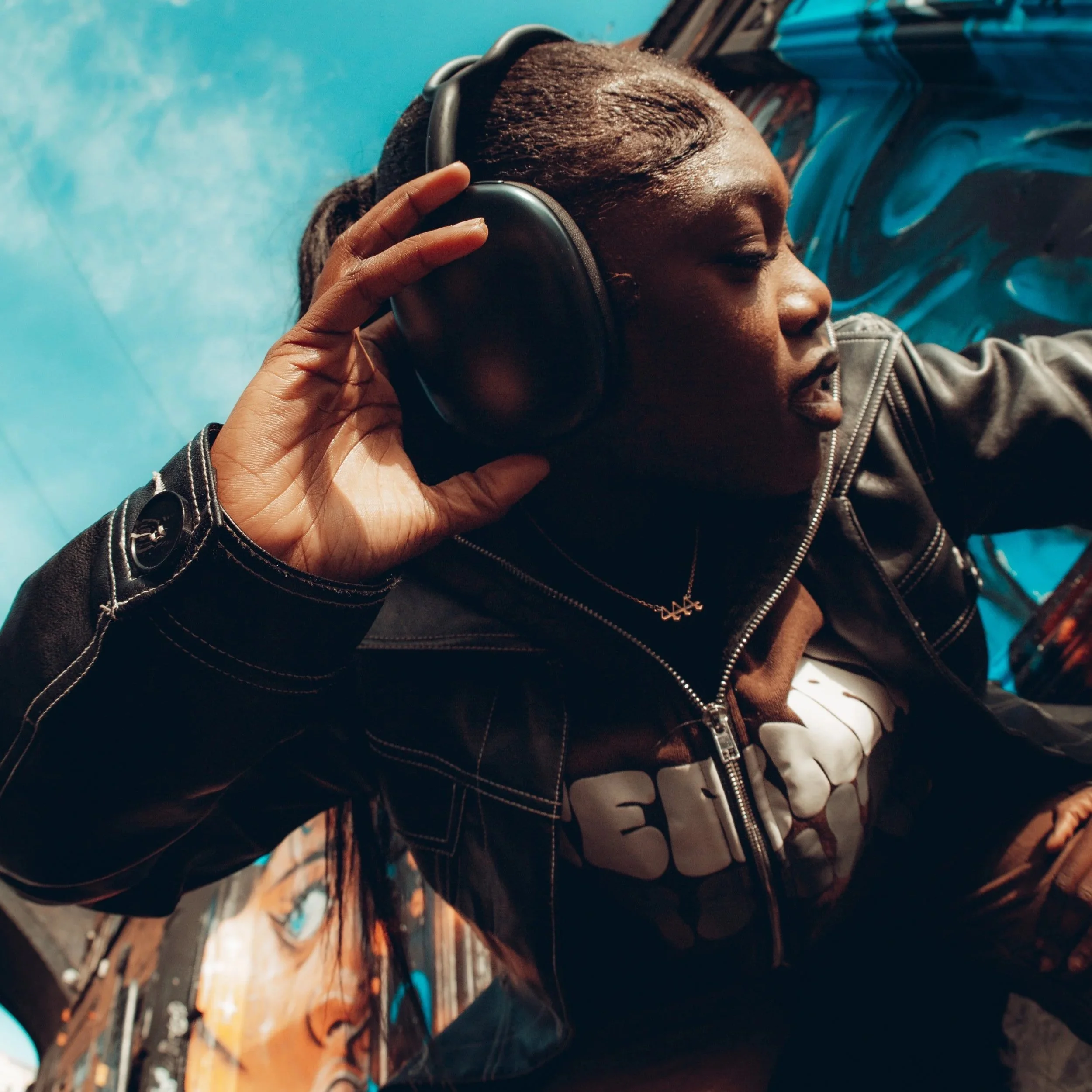 A woman with braided hair wearing a black leather jacket and headphones, stylized graffiti wall background.