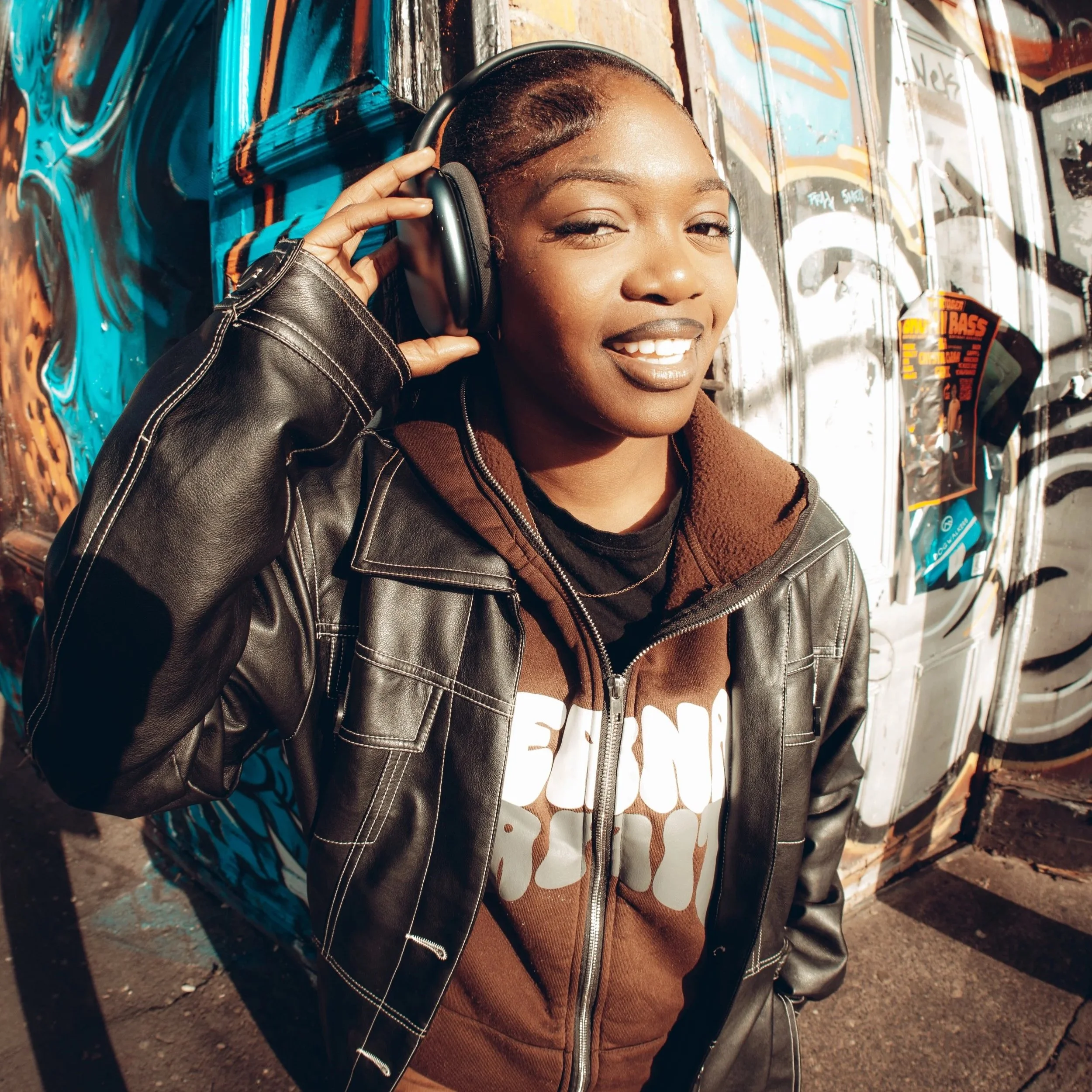A young woman with dark hair, wearing a black leather jacket and brown hoodie, smiles while holding headphones to her ear in front of a graffiti-covered wall.