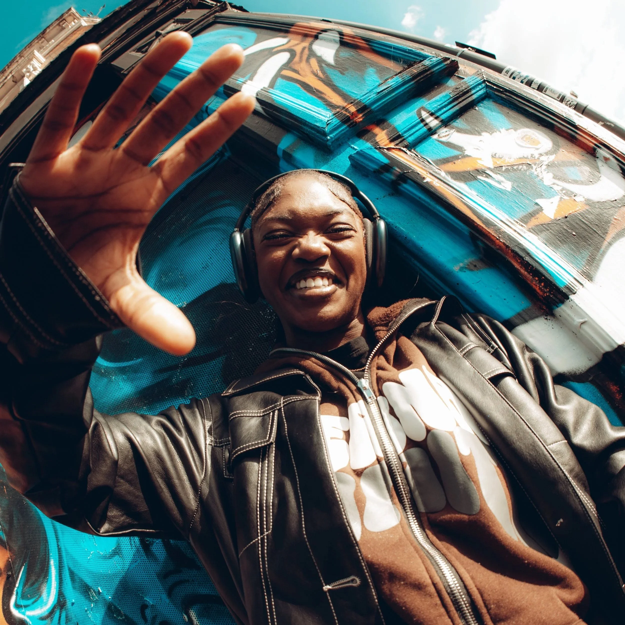A young person with short hair, wearing a leather jacket, a hoodie with a floral pattern, and headphones, smiling and waving while lying on a graffiti-covered surface outdoors, with a blue sky visible in the background.