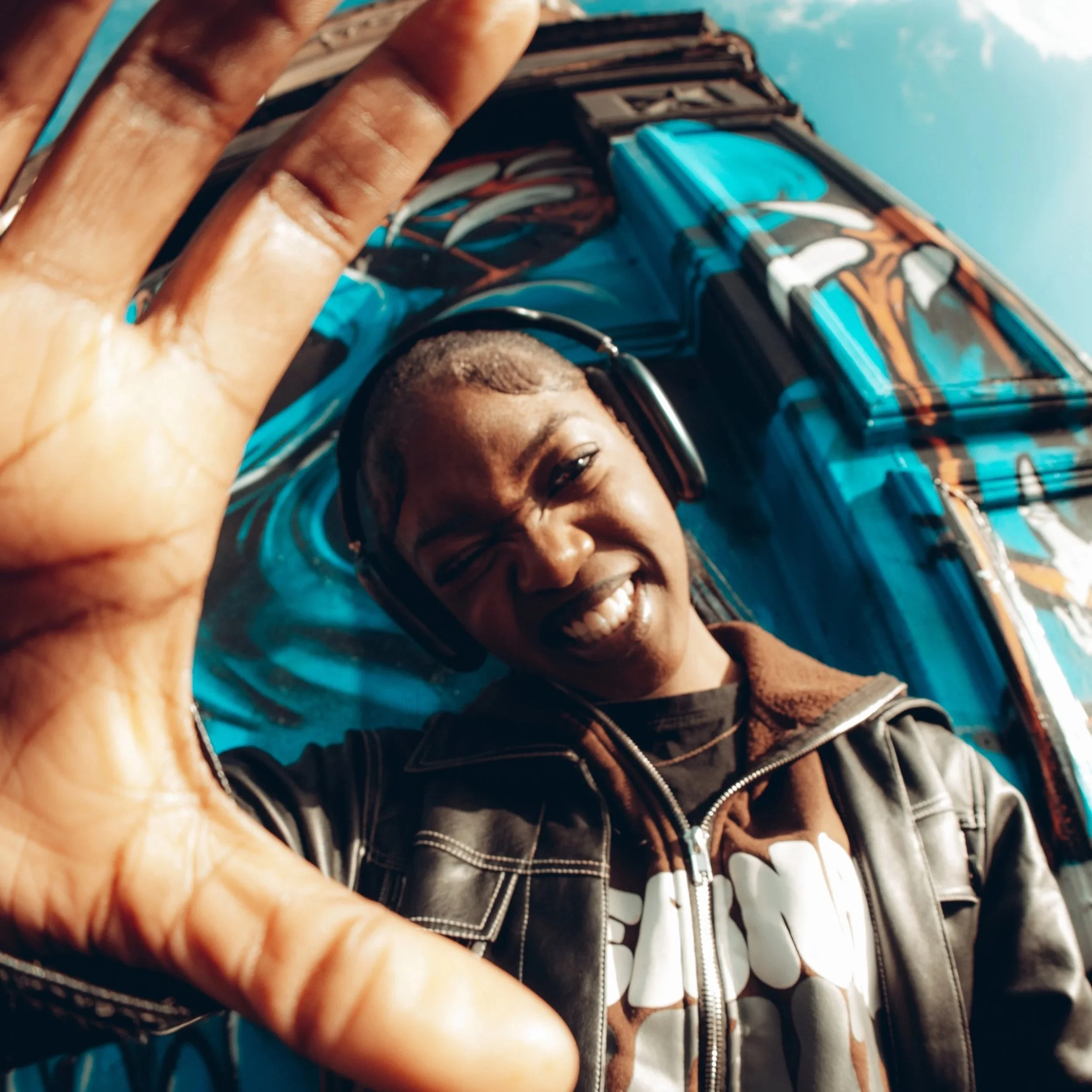A person smiling and waving while wearing headphones in front of a vibrant blue and orange painted wall.