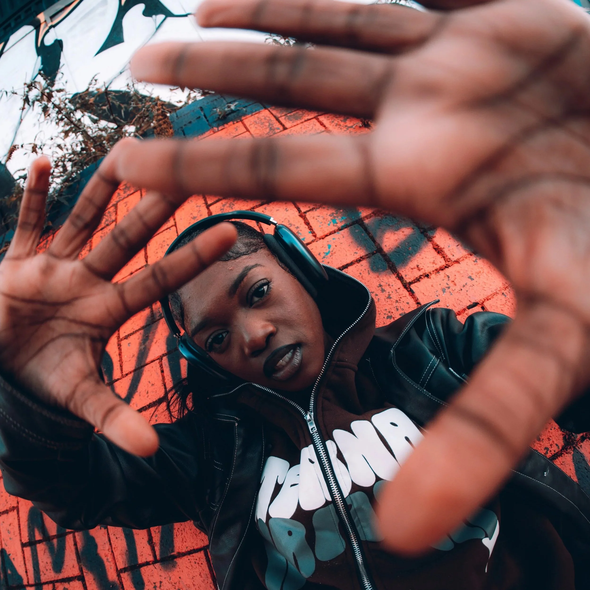 A woman wearing headphones and a black leather jacket with white text, lying on a red brick surface with graffiti, looking at the camera and reaching out with her hands