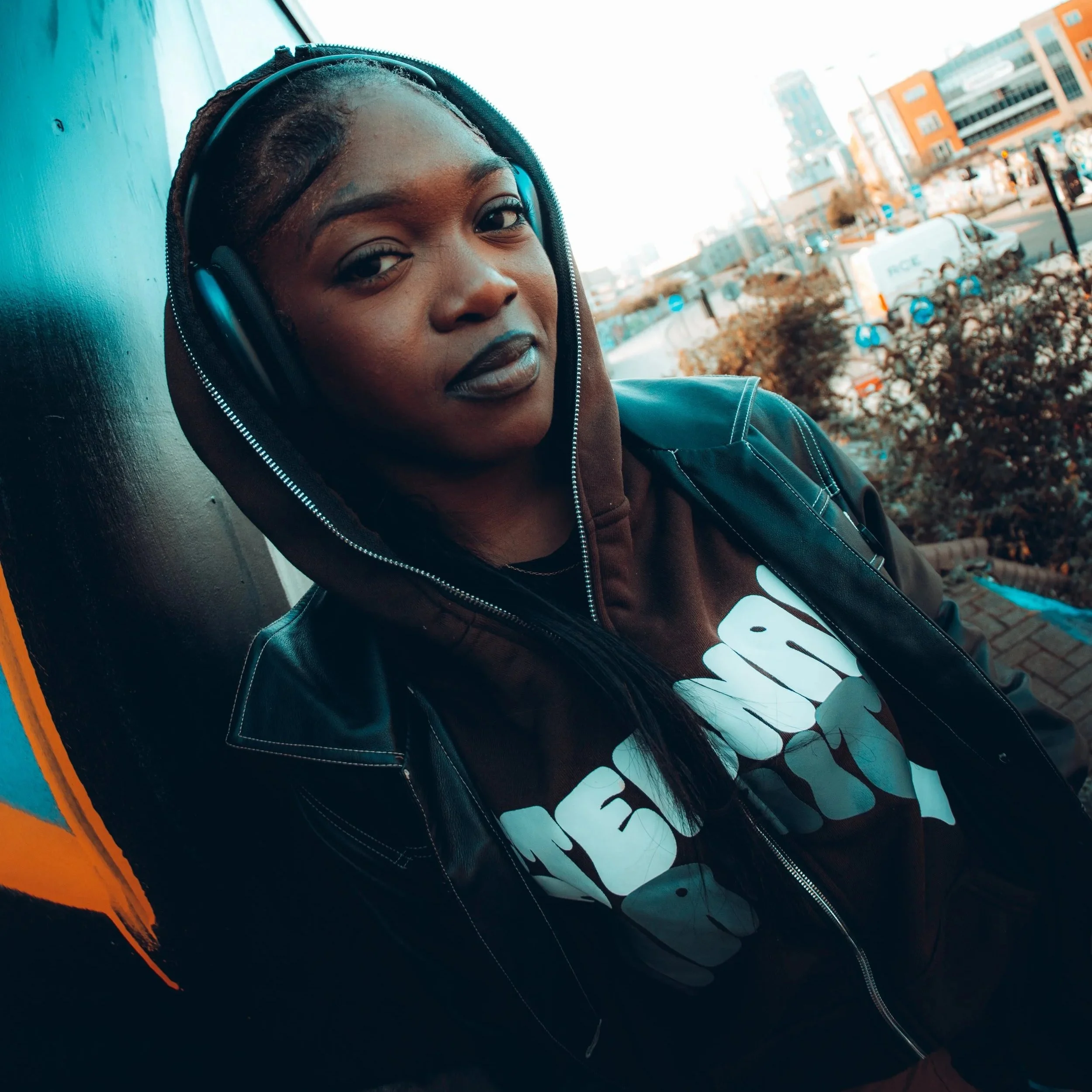 A young woman with dark skin and black braided hair wearing a black hoodie and headphones, standing outdoors near an urban street with modern buildings and vehicles in the background.