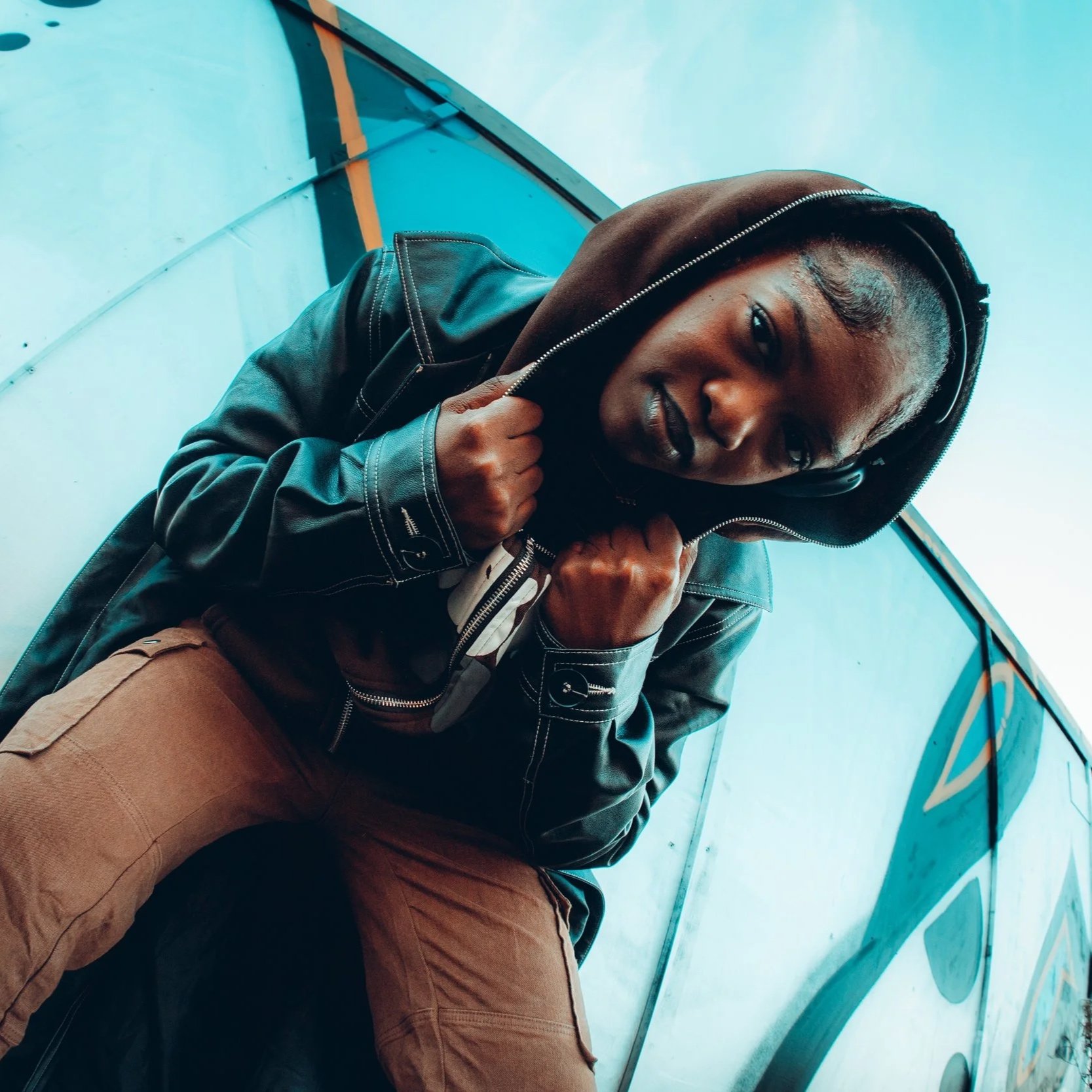 A person with braided hair, wearing a black leather jacket and brown pants, sitting against a colorful graffiti wall, looking directly at the camera.