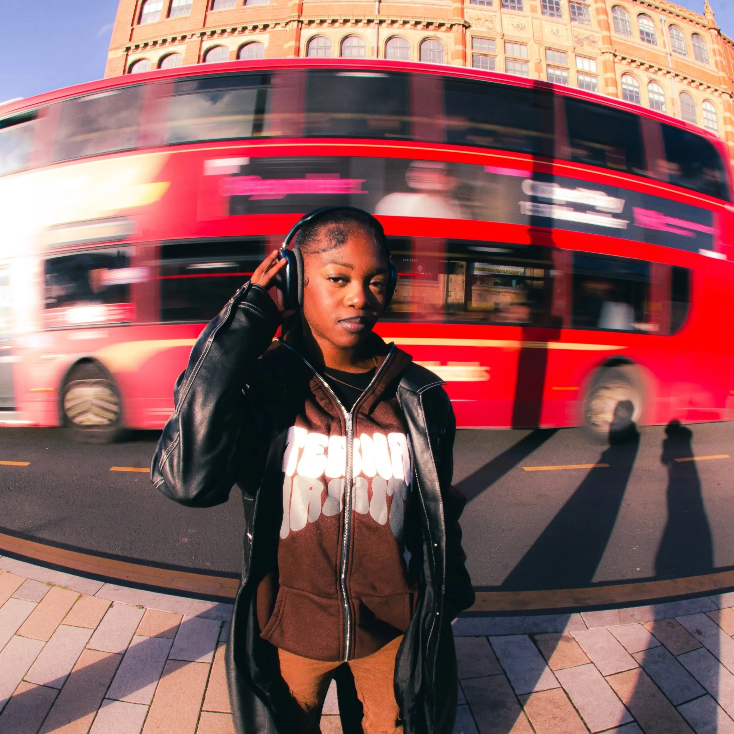 A young woman in a brown hoodie and black leather jacket stands on a city street, listening to music with headphones, with a red double-decker bus and historic buildings in the background.