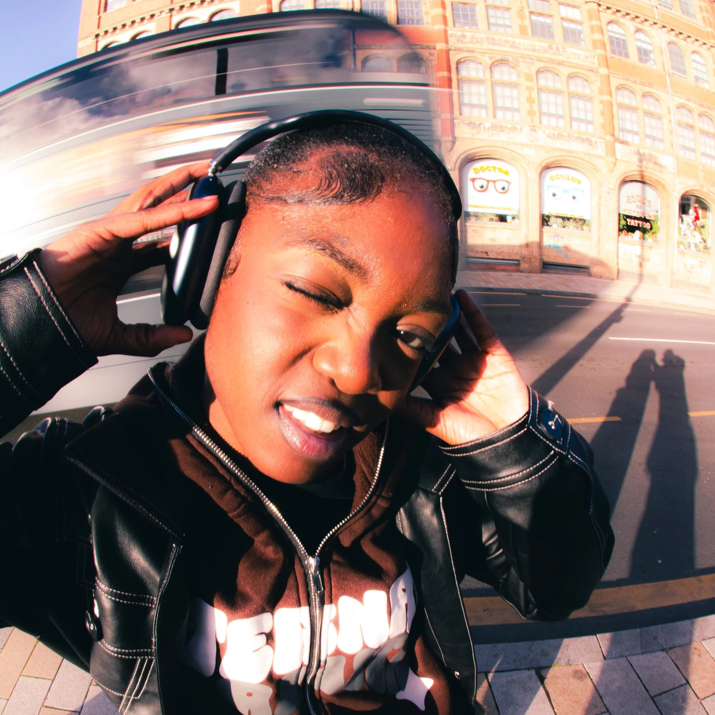 A young woman with short hair wearing headphones and a leather jacket takes a selfie on the street with a building and a moving bus in the background.