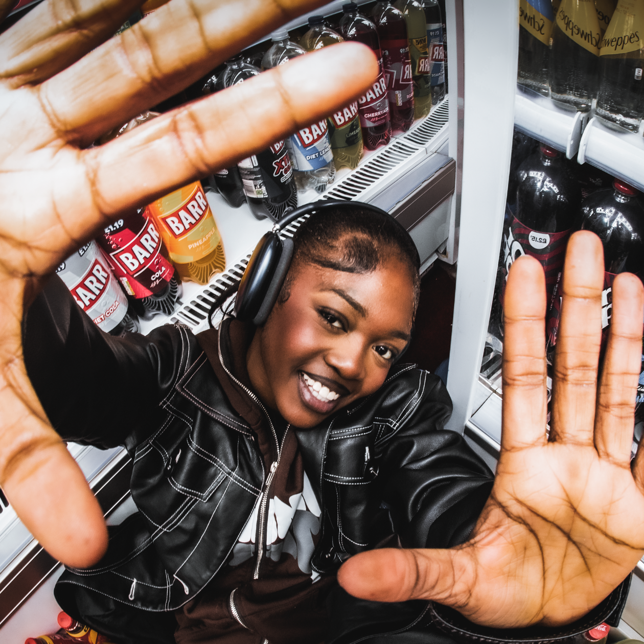 A woman with short hair, wearing a black leather jacket and headphones, smiling and holding her hands up in front of her in a friendly gesture in a store aisle with bottled beverages.