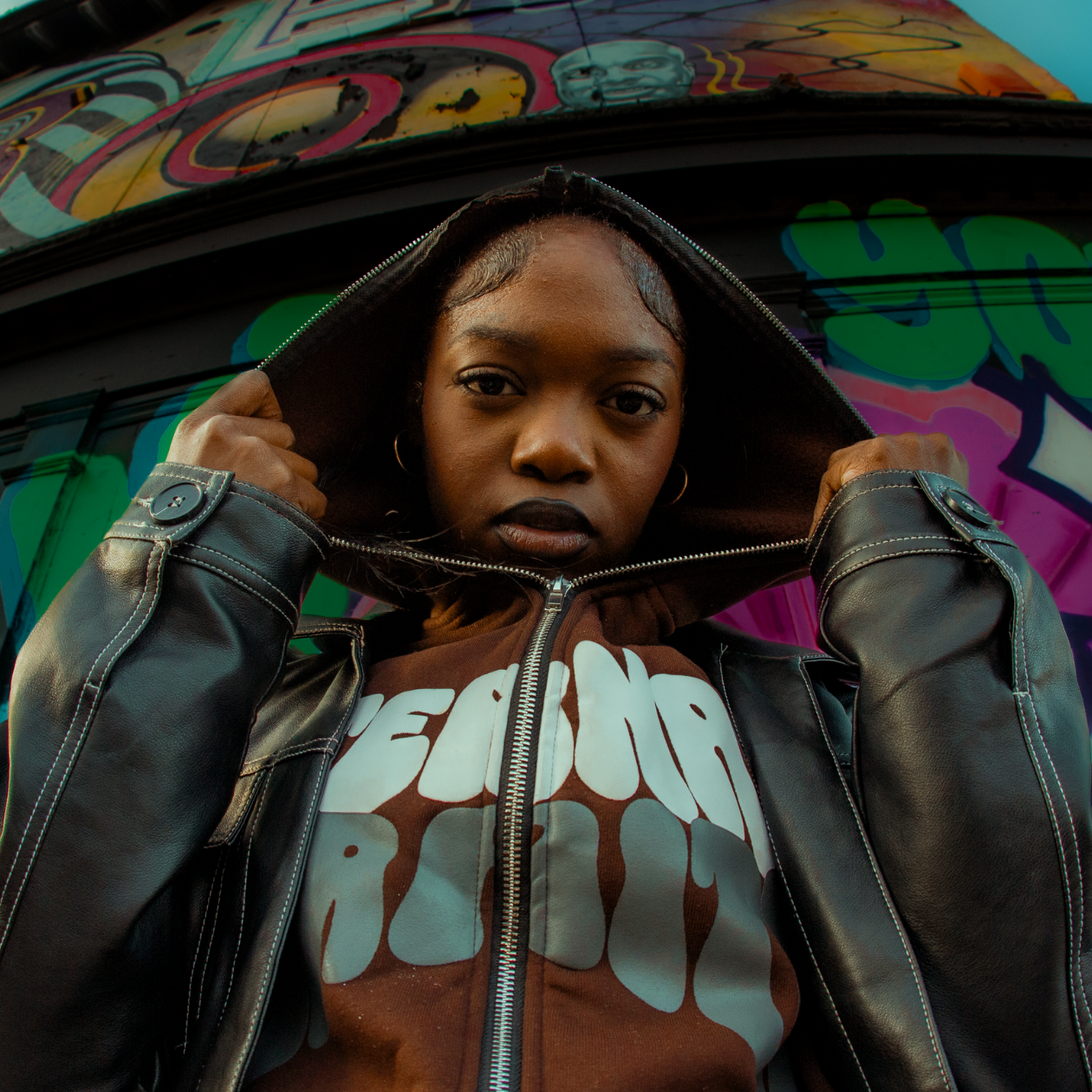 A woman with dark skin and black hair, wearing a brown hoodie with white graffiti-style text and a black leather jacket, looks directly at the camera while pulling the hoodie over her head. She is standing in front of a colorful graffiti mural.