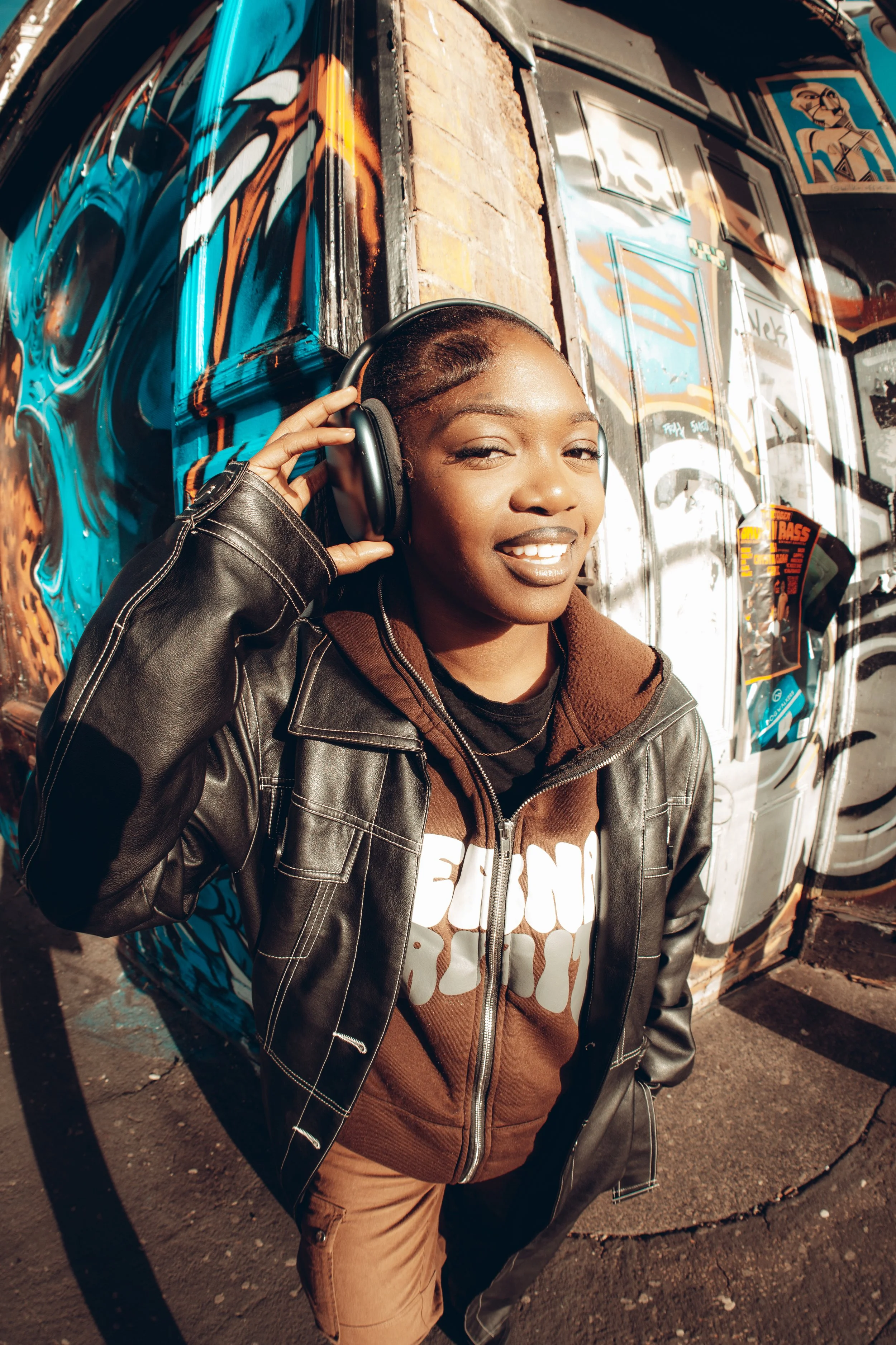 A young woman in a leather jacket and brown hoodie listens to music with headphones outside against a graffiti-covered wall.